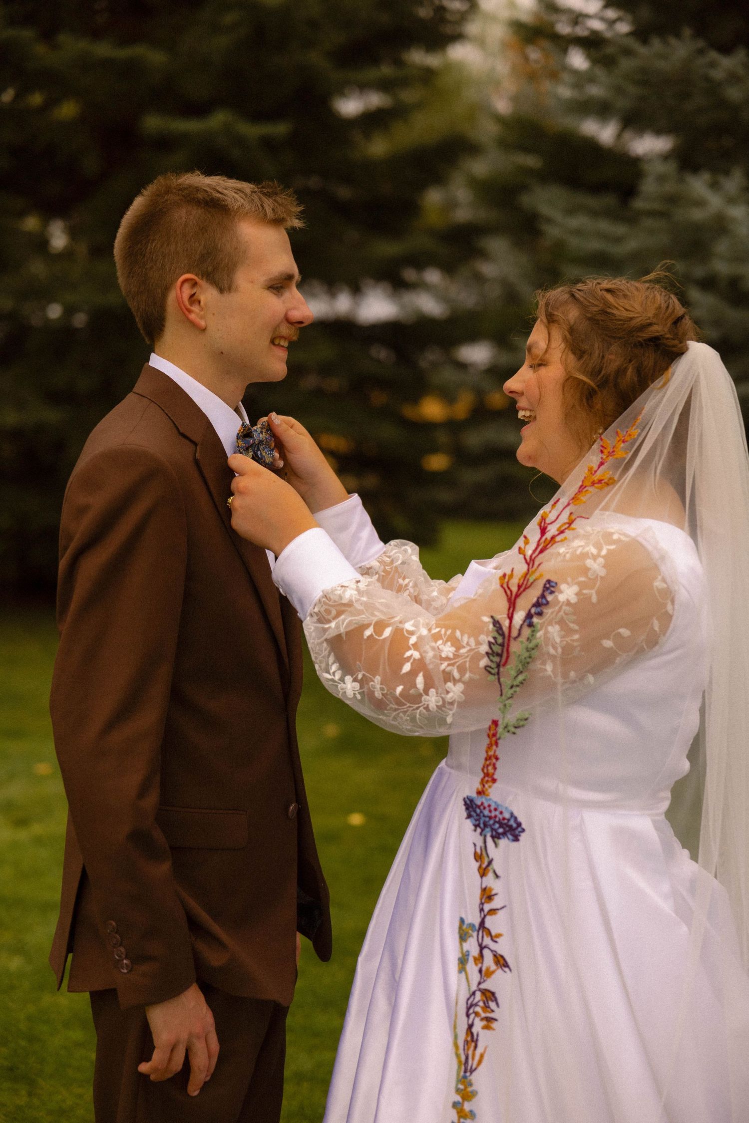 Bride in embroidered white gown adjusts groom's boutonniere outdoors, both smiling joyfully on their wedding day.