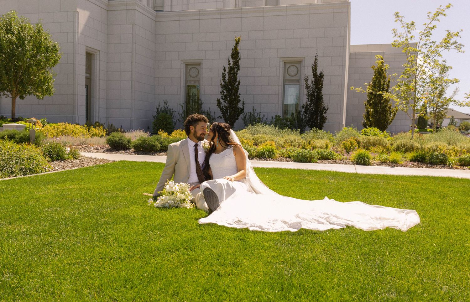 Newlywed couple sharing a kiss while sitting on green grass outside a white wedding temple on their special day.