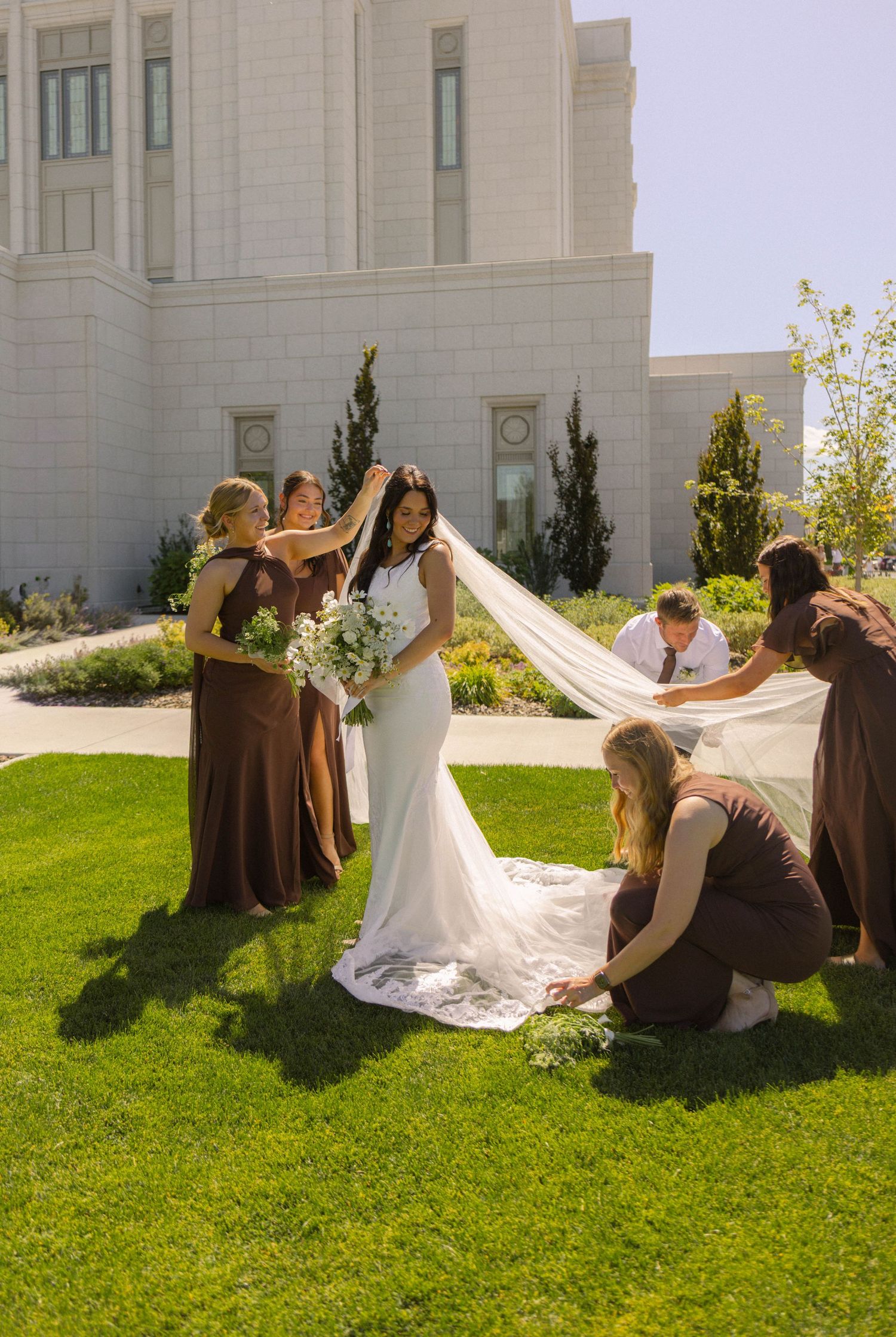 Bridesmaids in brown dresses arrange bride's long veil on green lawn outside a white LDS temple on sunny wedding day.