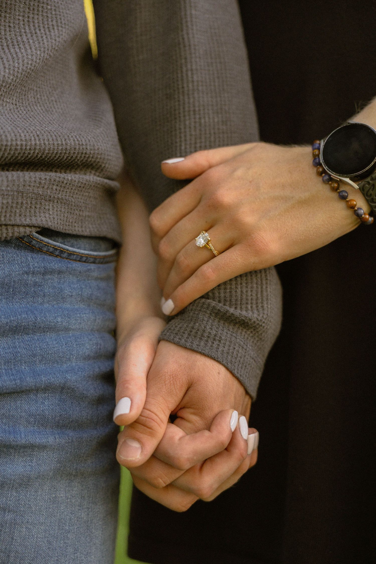 Couple holding hands with a diamond engagement ring on gold band, white manicured nails, smart watch, and beaded bracelet.