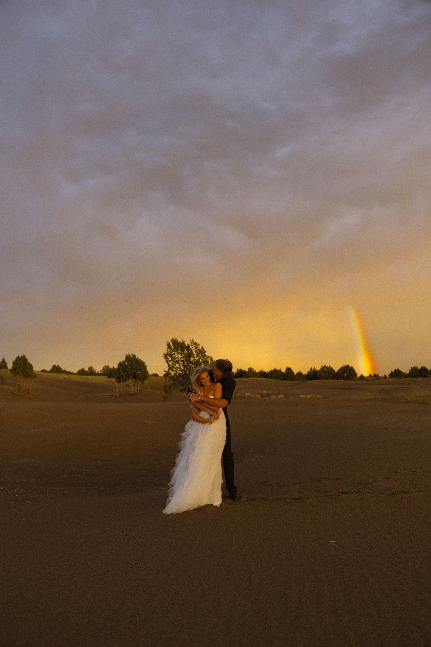 Newlywed couple embracing on sandy dunes at sunset with a rainbow in the dramatic cloudy sky behind them.