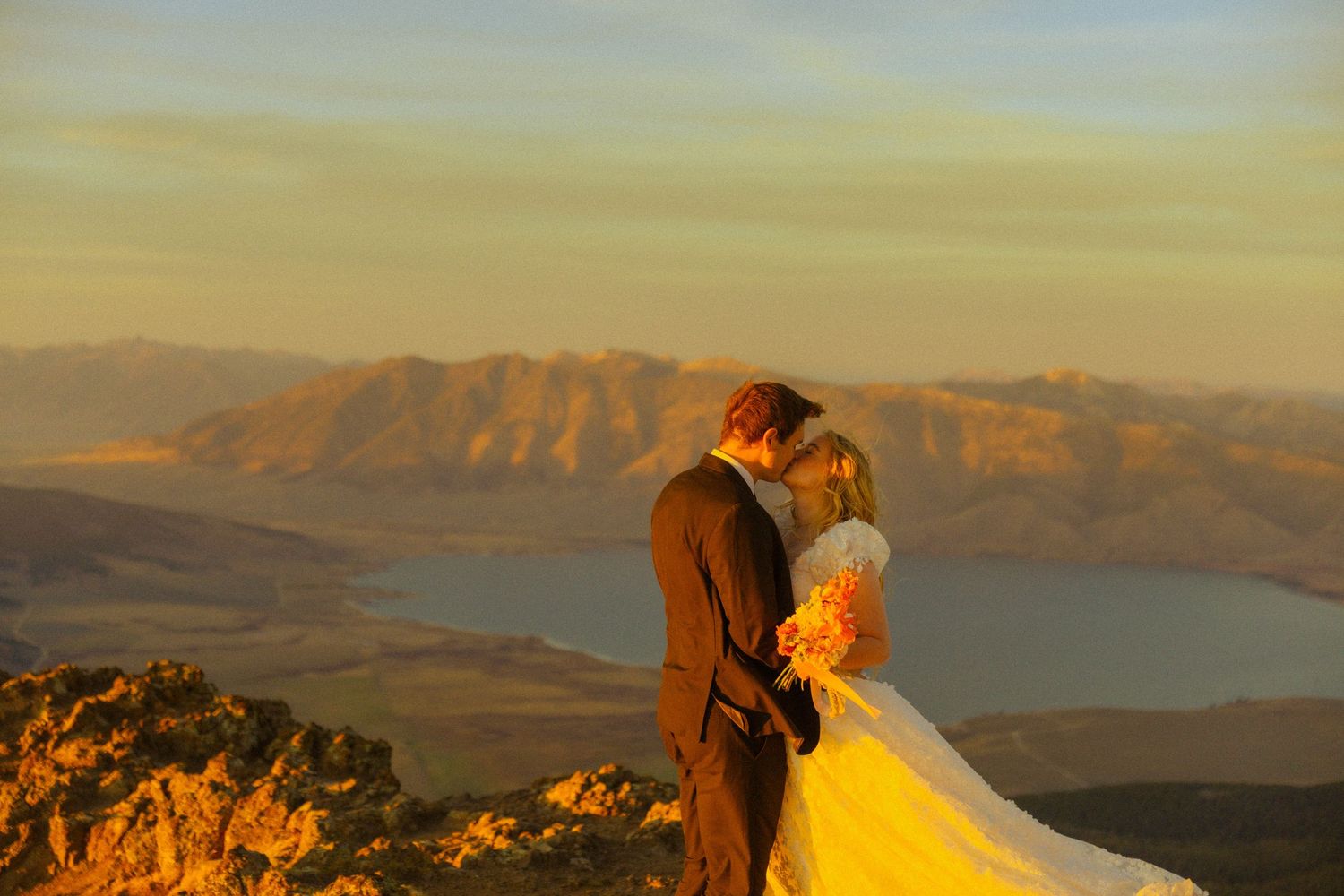 Bride and groom kissing on a mountain summit at golden hour with scenic lake and mountain range backdrop.