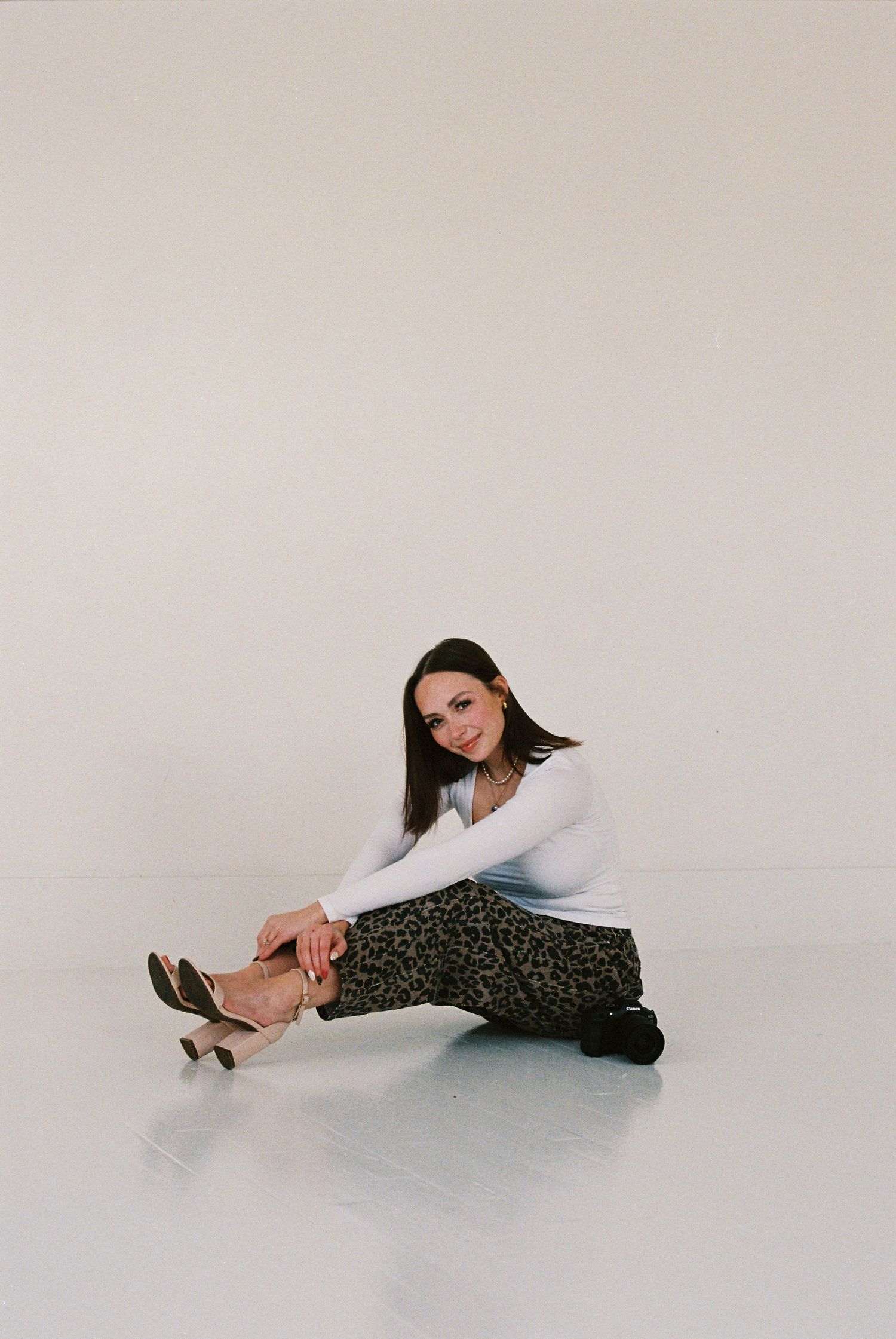 Woman in white top and leopard print pants sits on floor smiling, wearing heeled boots in a bright studio.