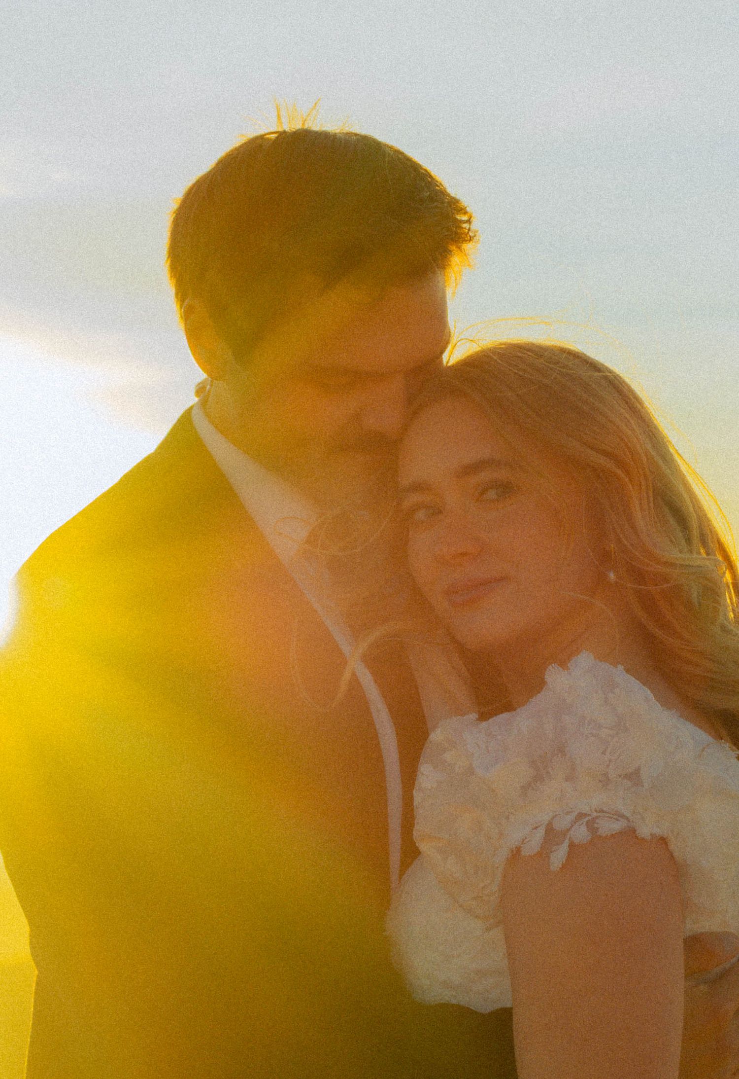 Couple in wedding attire embracing at golden hour sunset, warm glowing light surrounding them romantically.
