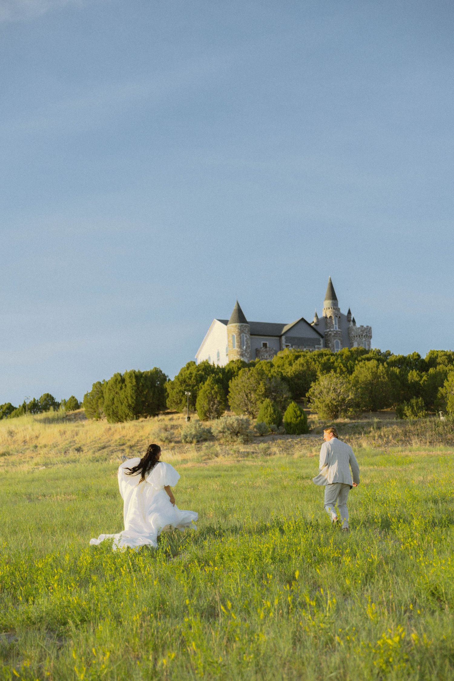 Bride and groom walking toward a castle on a hill surrounded by green fields and trees under a blue sky.