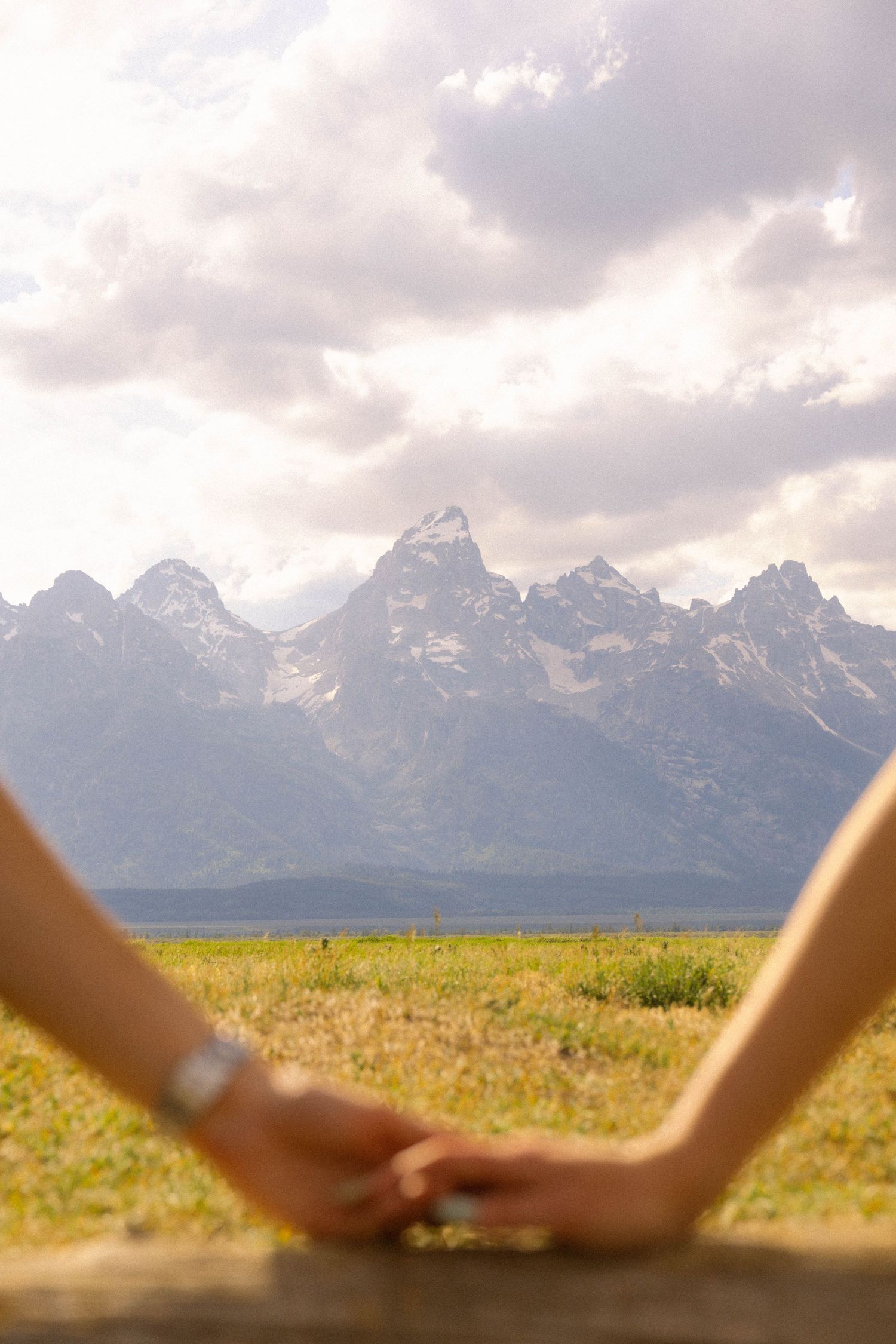 Couple holding hands on a wooden fence with Grand Teton mountain range and green meadow in background.