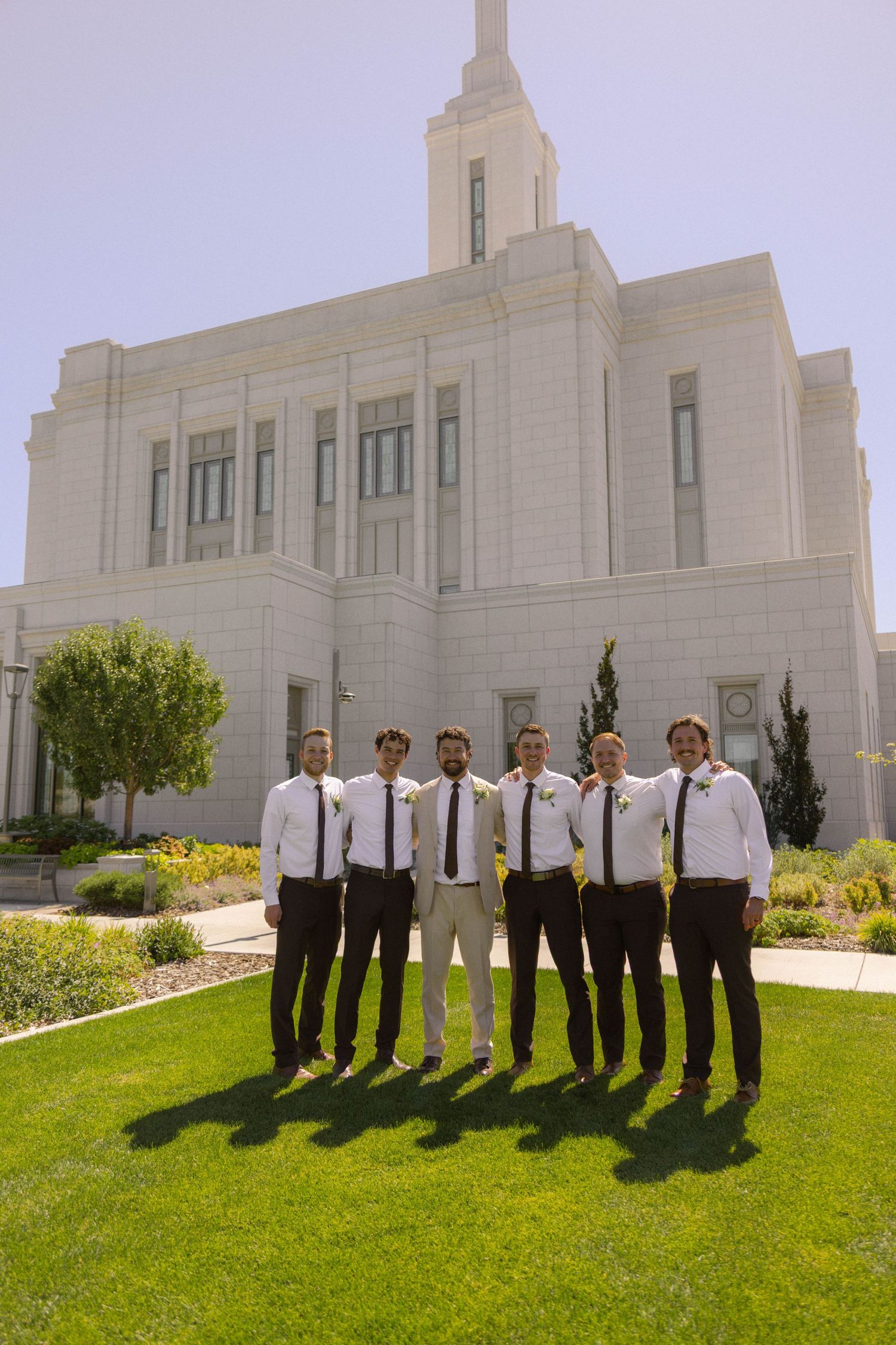Groom and groomsmen in white shirts and dark ties pose on green lawn before a white LDS temple on a sunny day.