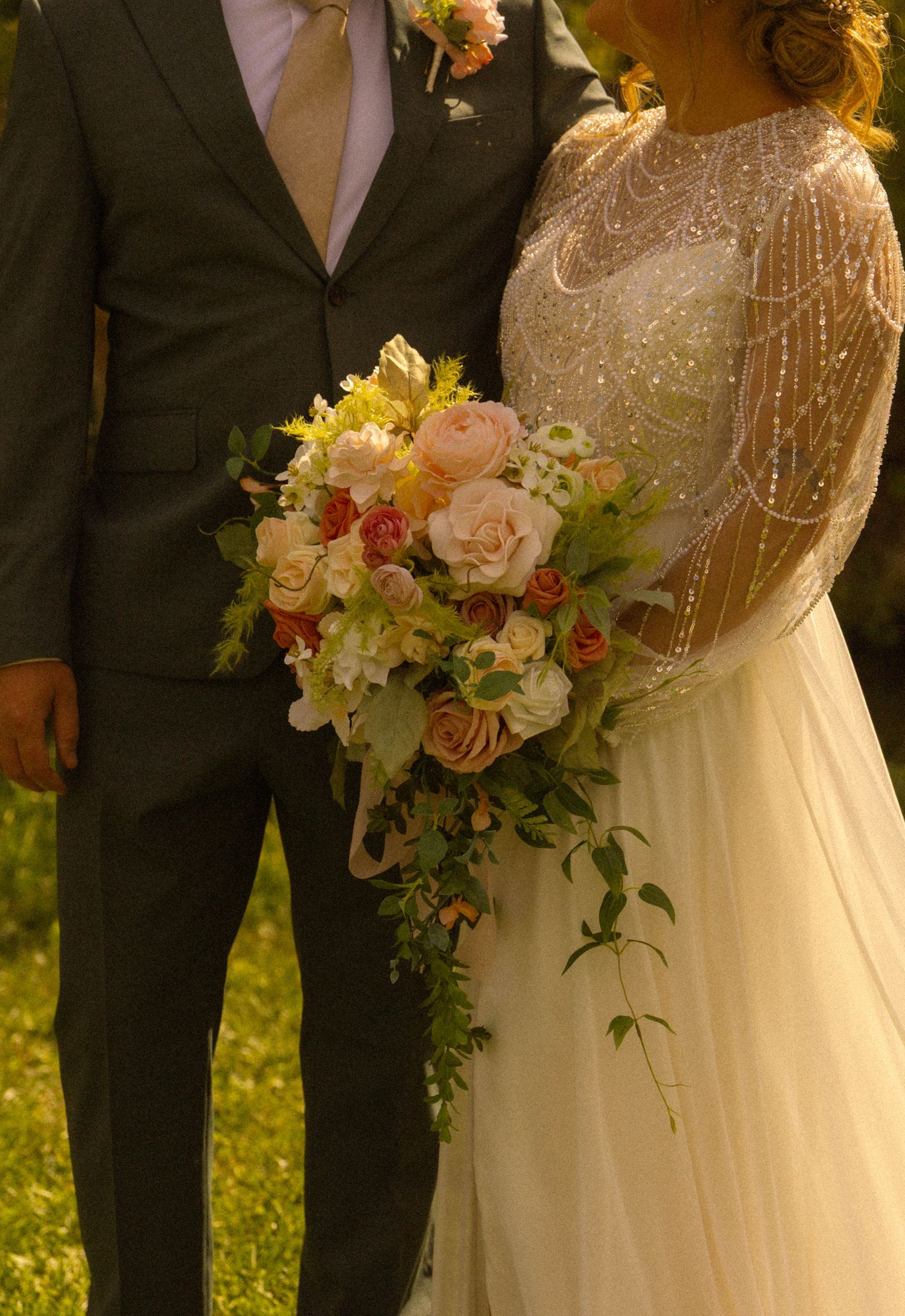 Bride in beaded gown holding cascading peach and pink rose bouquet standing beside groom in gray suit outdoors.