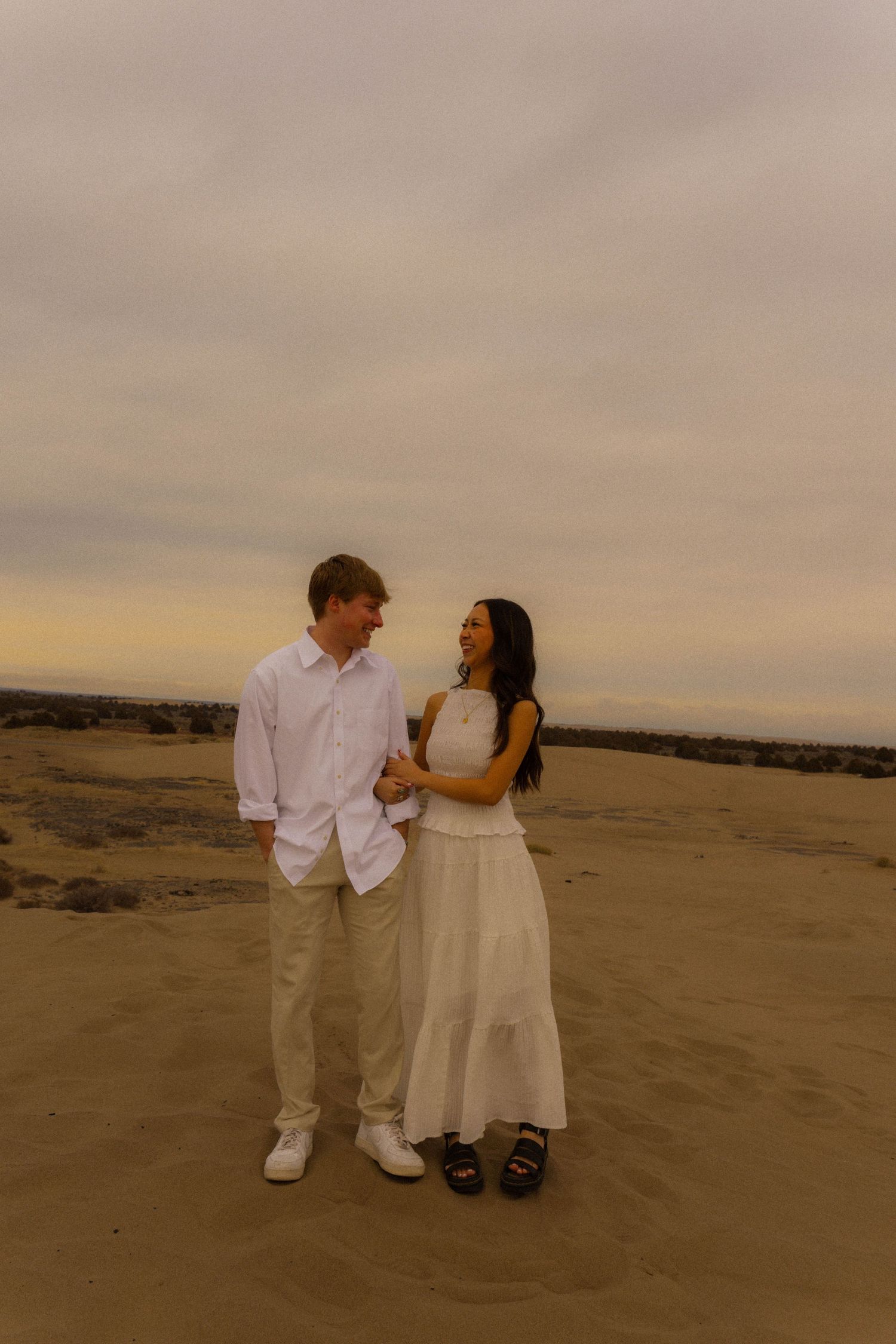 Couple in white outfits holding hands on sandy desert dunes at sunset, smiling at each other romantically.