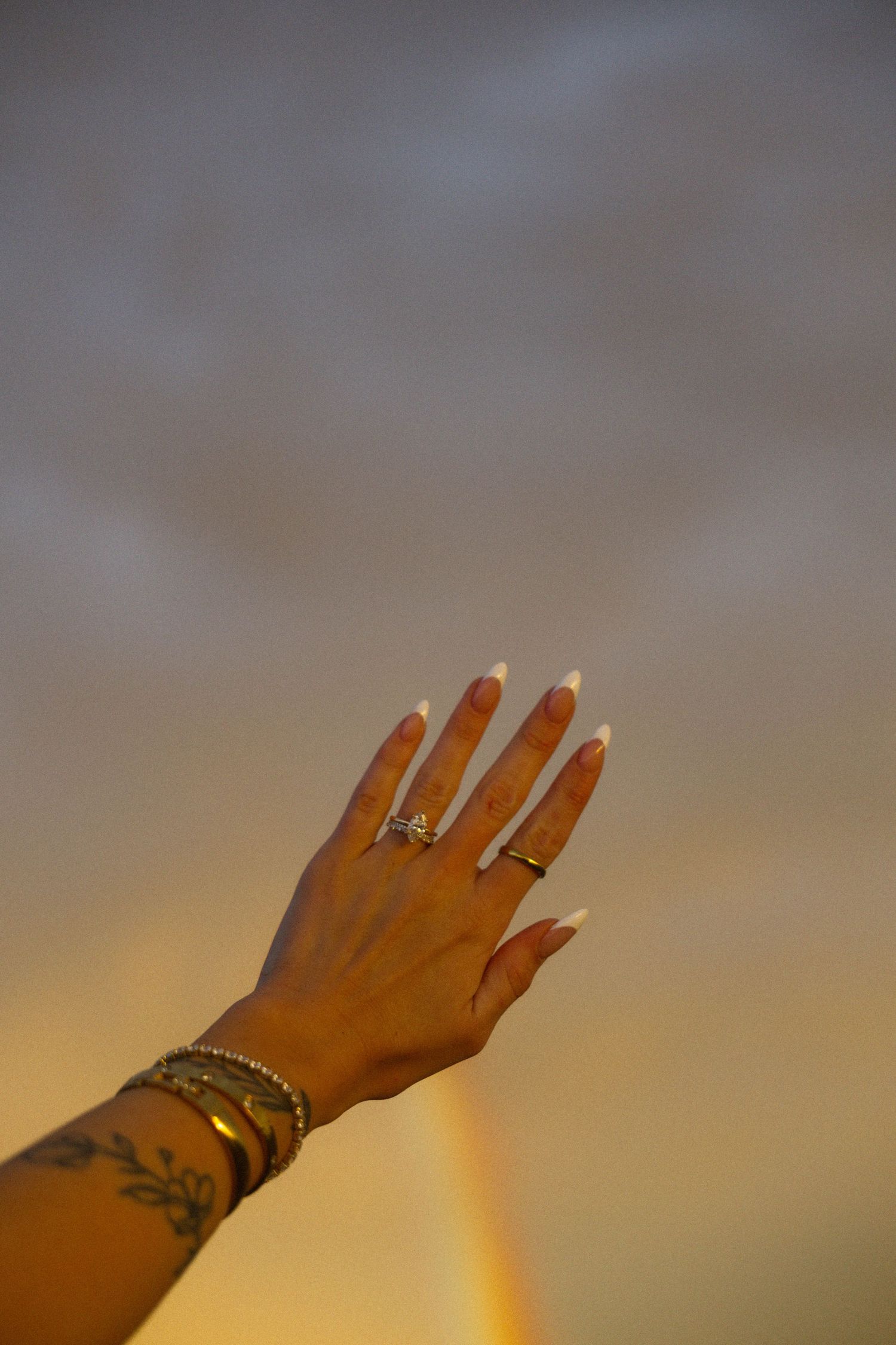 A woman's hand with white French tip nails, gold rings, bracelets, and a floral wrist tattoo raised against a rainbow sky.