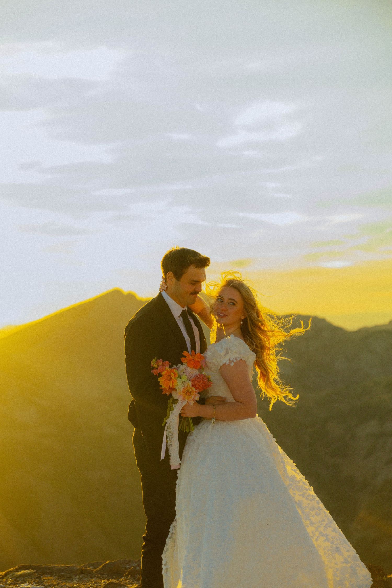 Bride and groom embrace at golden sunset on mountain peak, bride holding colorful bouquet in white lace gown.