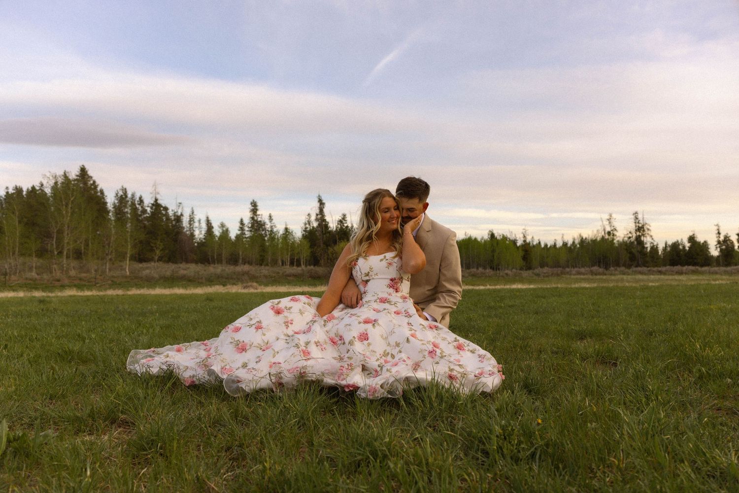Couple in floral wedding dress sitting together on green meadow with forest backdrop at sunset.