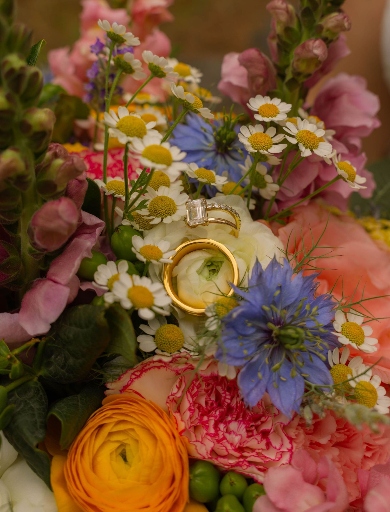 Gold wedding bands rest on a vibrant bridal bouquet with daisies, blue nigella, pink carnations, and yellow ranunculus flowers.