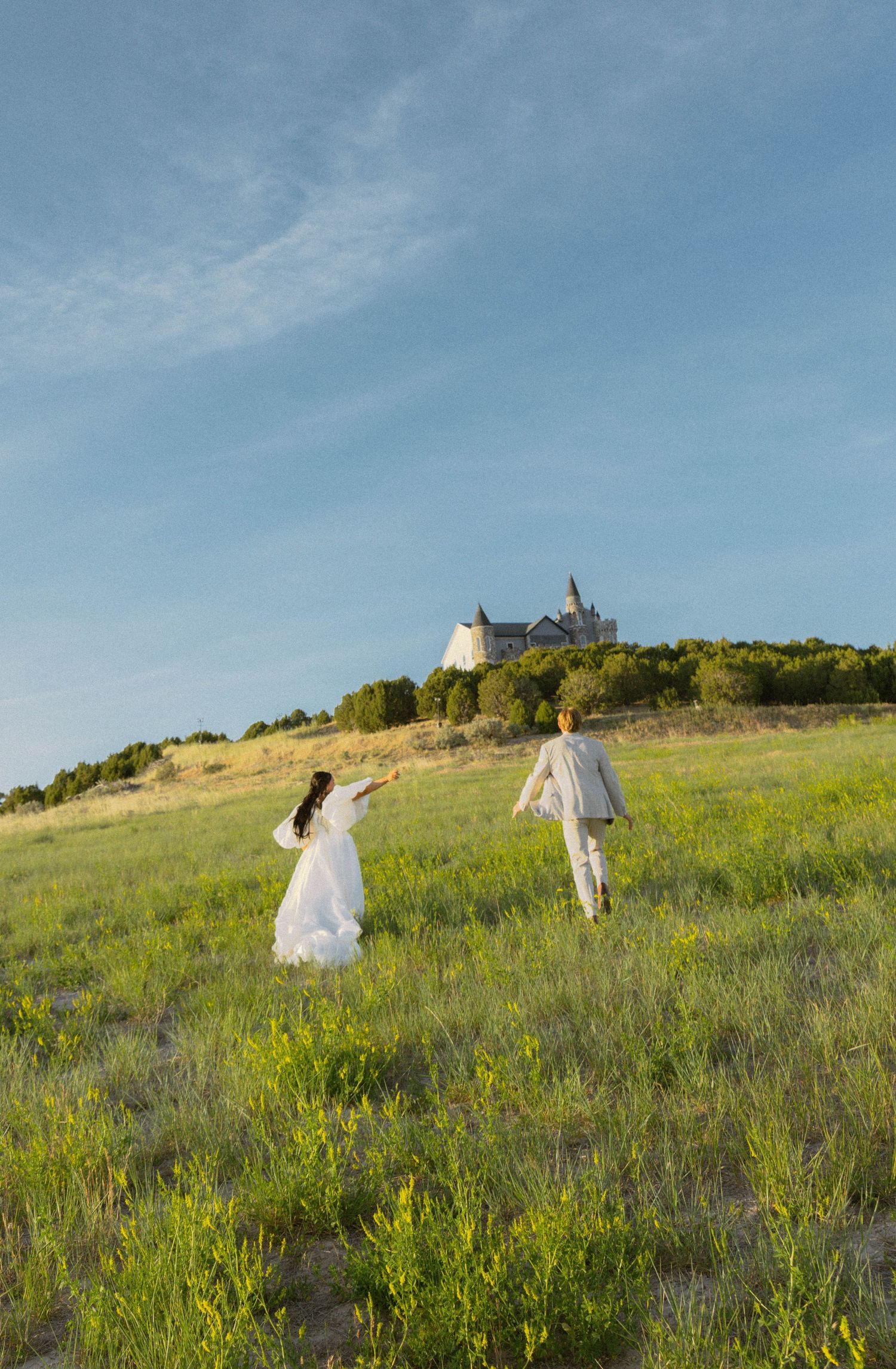 A bride and groom run through a green meadow toward a castle on a hill under a blue sky.
