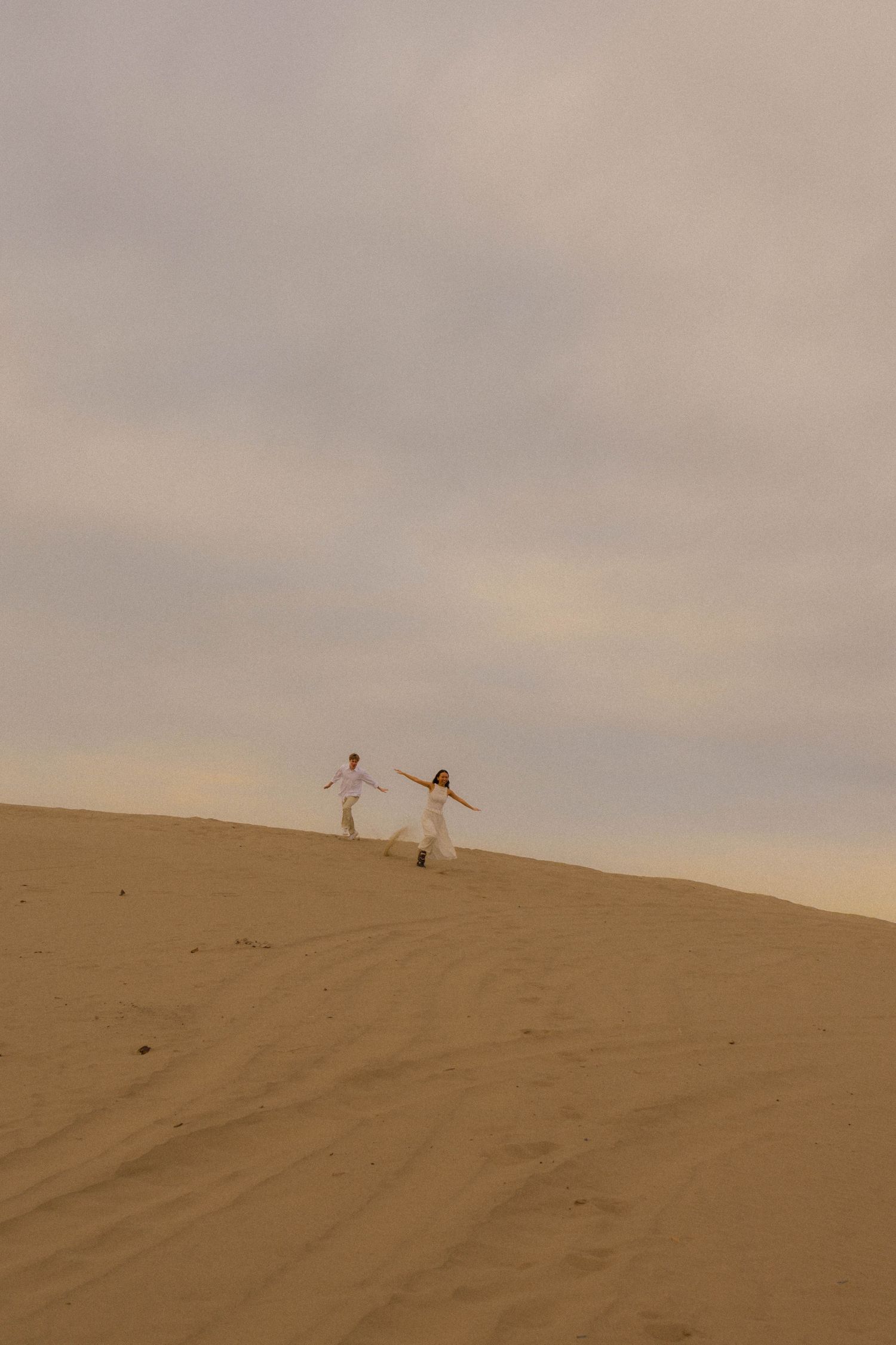 Couple in white outfits holding hands on a sandy desert dune under a cloudy sky.