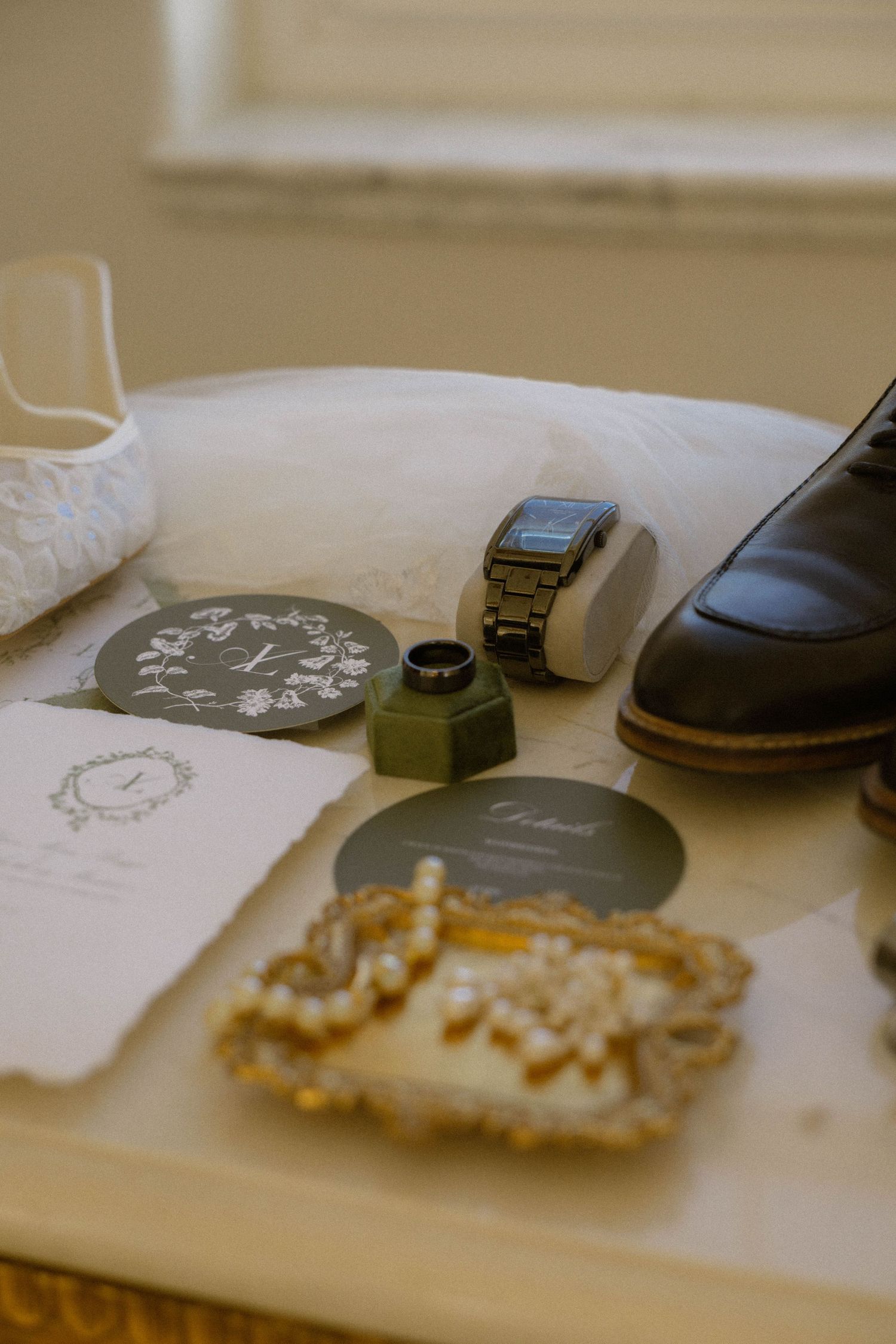 Wedding flat lay with rings, watch, invitation cards, pearl jewelry, dress shoes, and lace bridal accessories on a white surface.