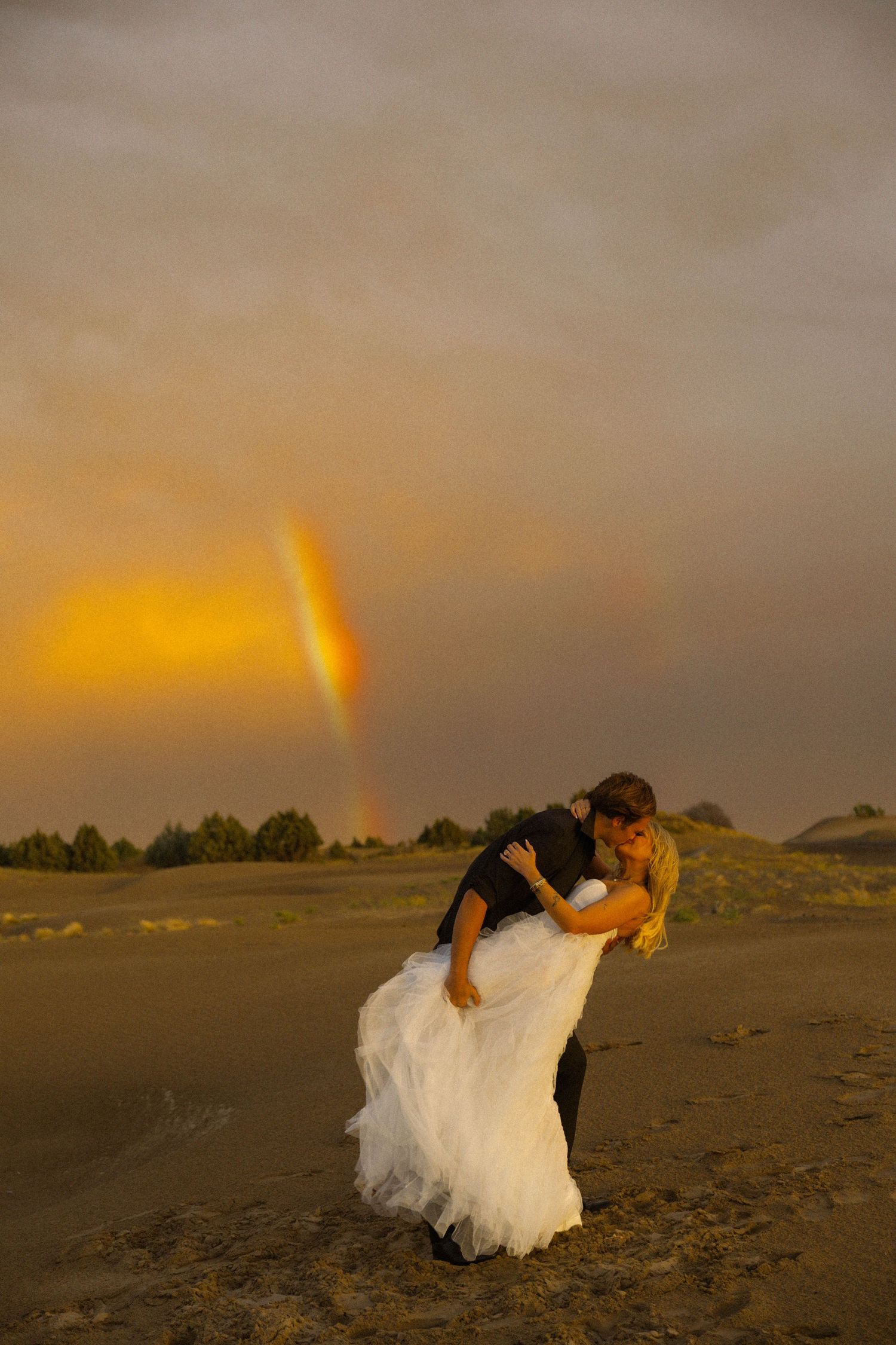 Bride and groom share a romantic dip kiss on a sandy beach at sunset with a stunning double rainbow in the background.