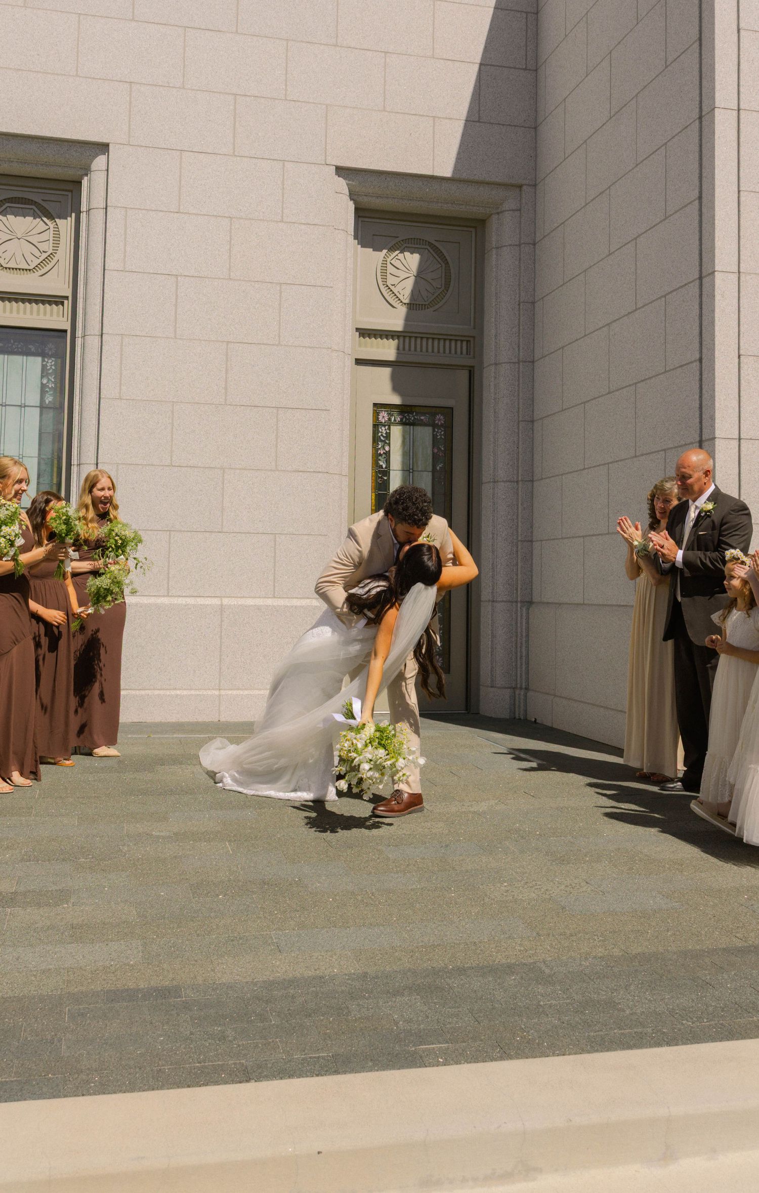 Bride and groom share a romantic dip kiss outside a wedding venue, surrounded by bridesmaids in brown dresses.