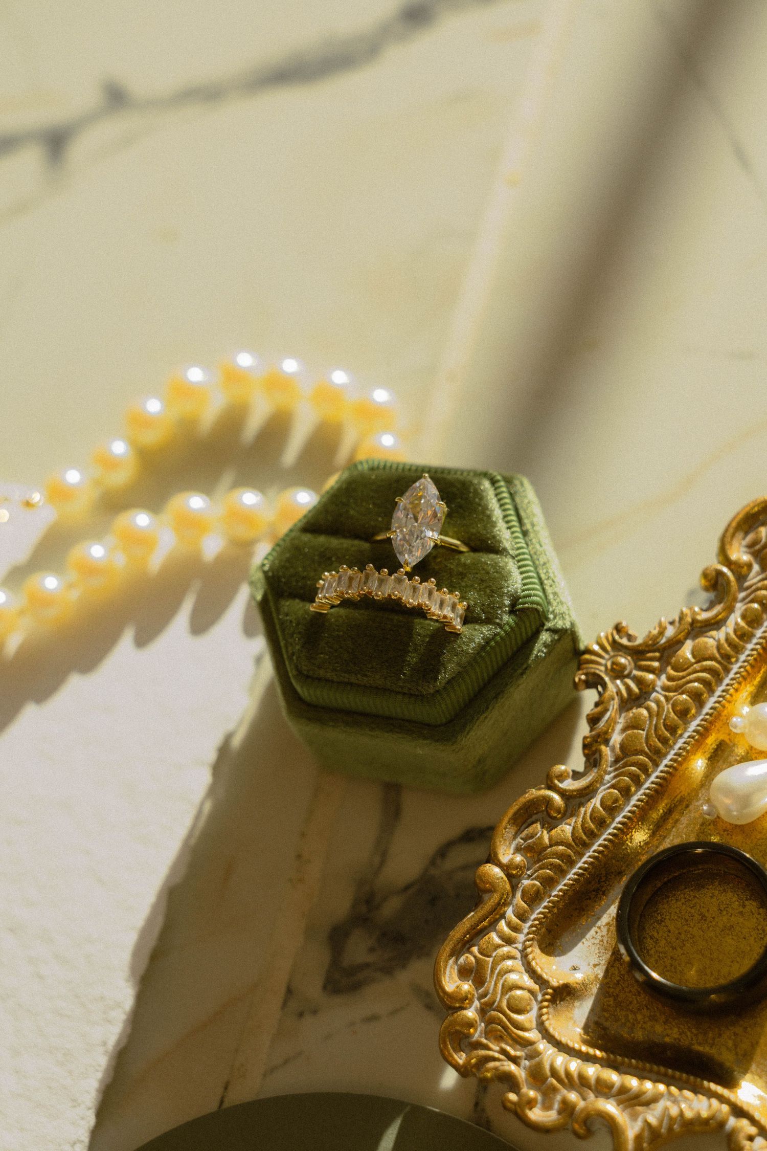 Diamond ring in green velvet box beside pearl necklace and gold ornate tray on marble surface.