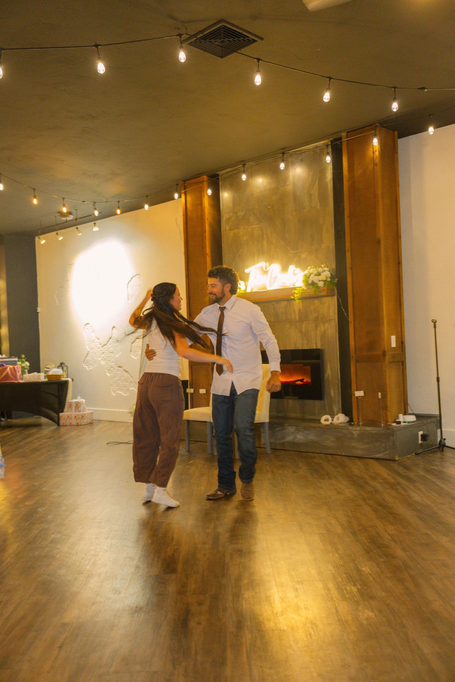 Couple dancing together in a warmly lit venue with string lights and a fireplace backdrop.