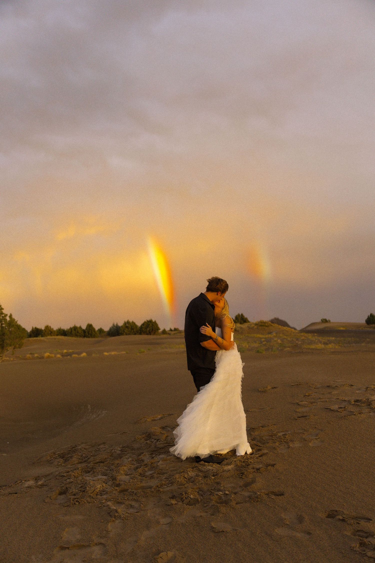Bride and groom kissing at sunset on sandy dunes with a double rainbow glowing in the dramatic cloudy sky.