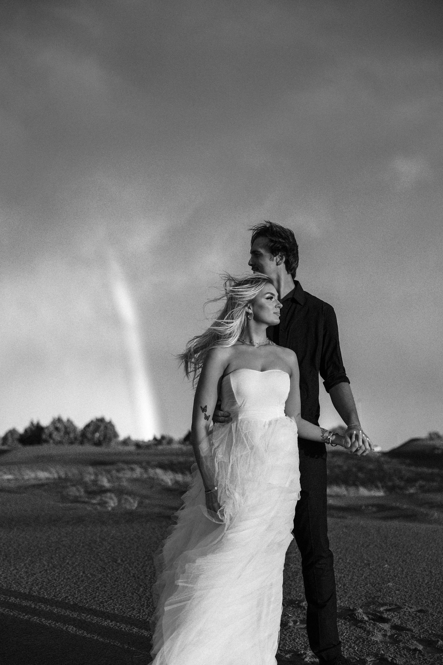 Black and white wedding photo of bride and groom holding hands on road with dramatic sky and rainbow behind them.