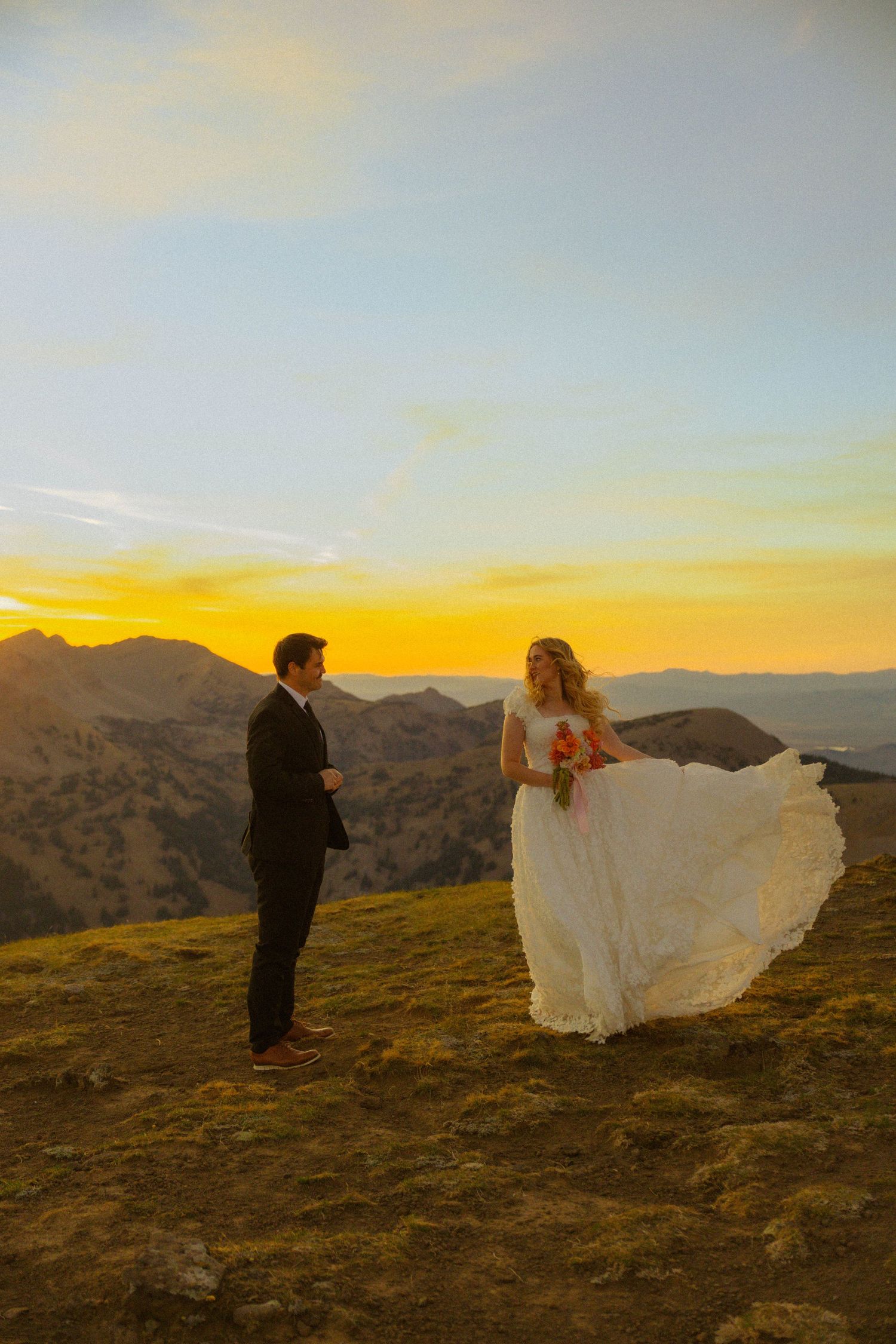 Bride and groom share a romantic sunset moment on a mountain hilltop, her white gown flowing in the breeze.