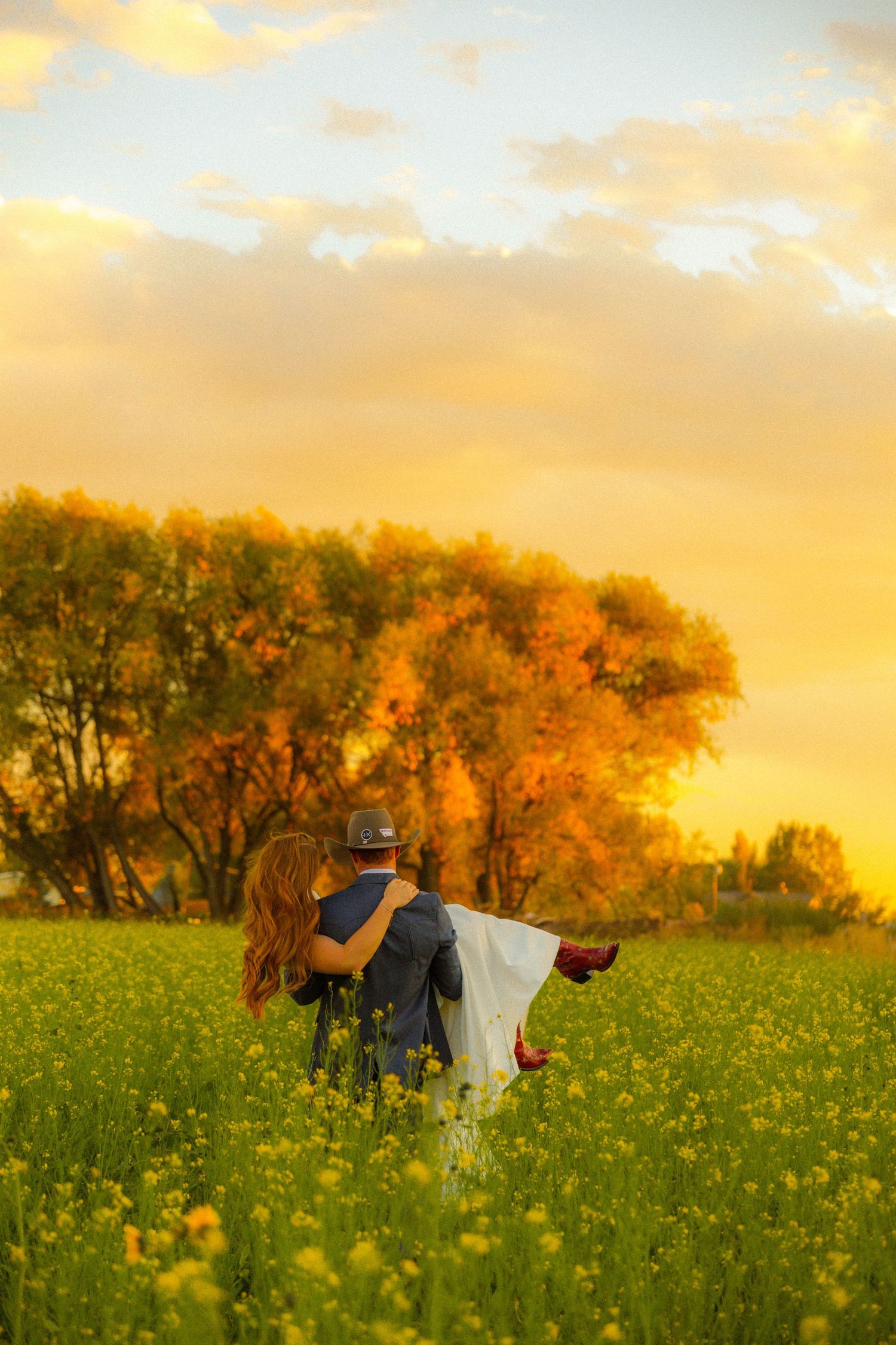 Couple embracing in a golden sunset field of yellow flowers with autumn trees in the background.