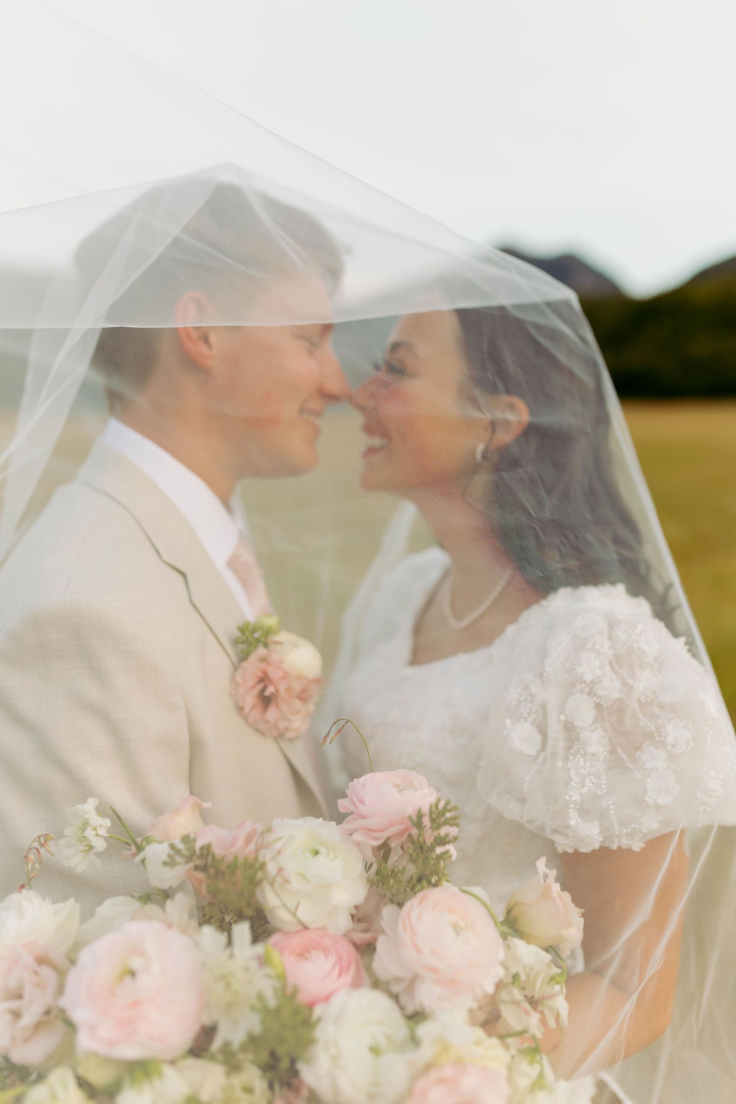 Bride and groom share a romantic moment under a sheer veil, with a soft pink floral bouquet in the foreground.