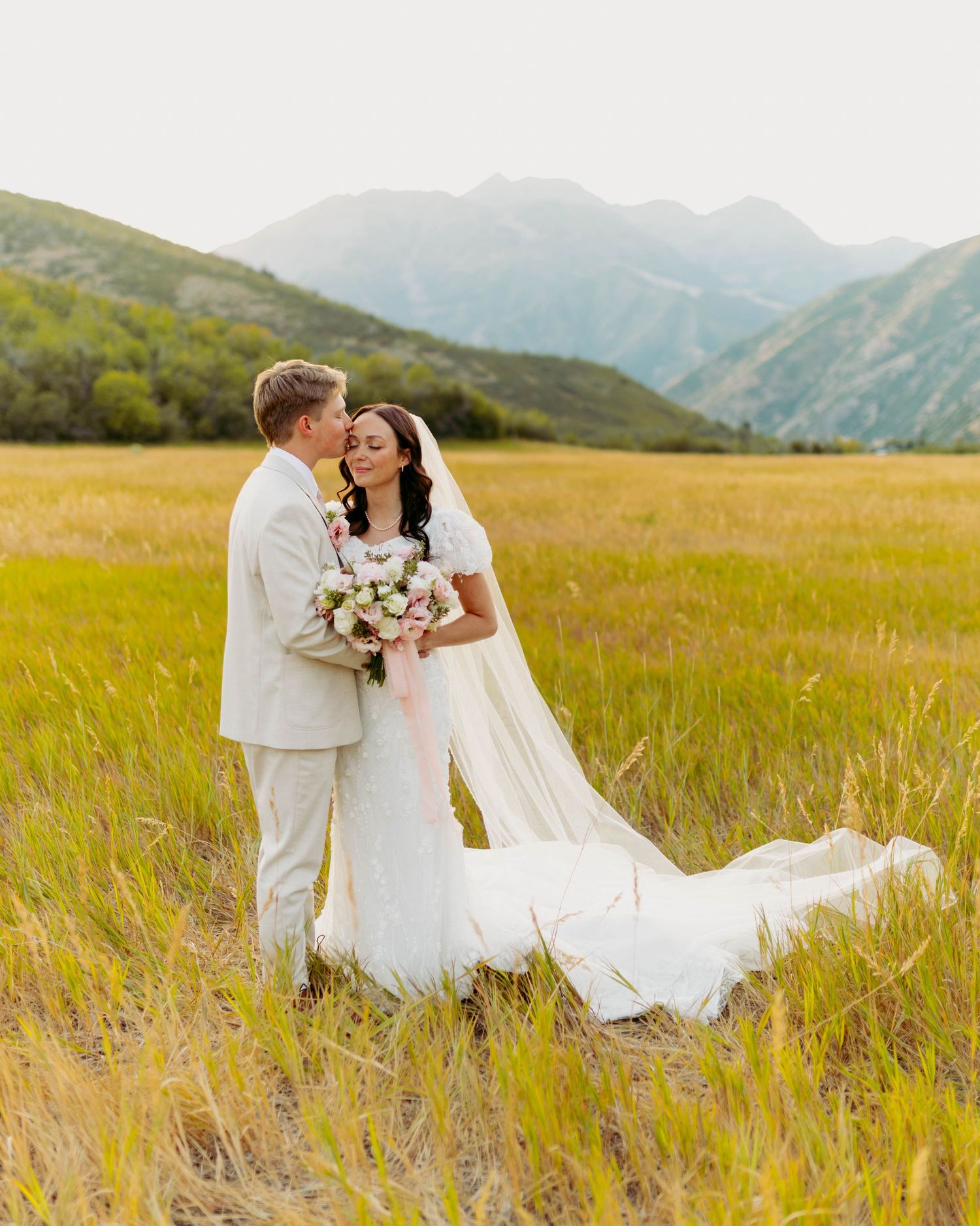 Bride and groom share a romantic moment in a golden meadow with stunning mountain backdrop on their wedding day.