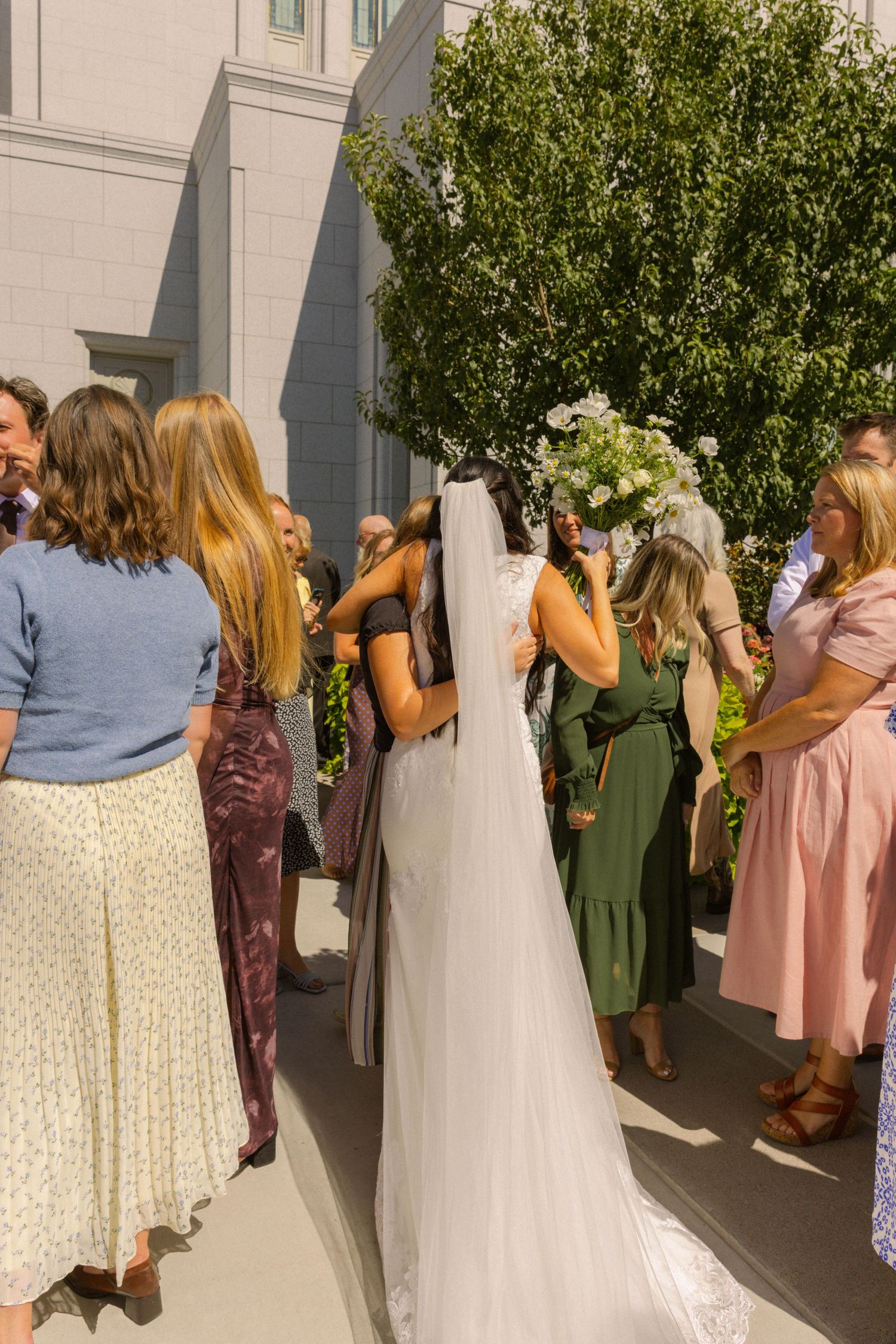 Bride in white gown and veil holding floral bouquet surrounded by wedding guests outdoors near a building.