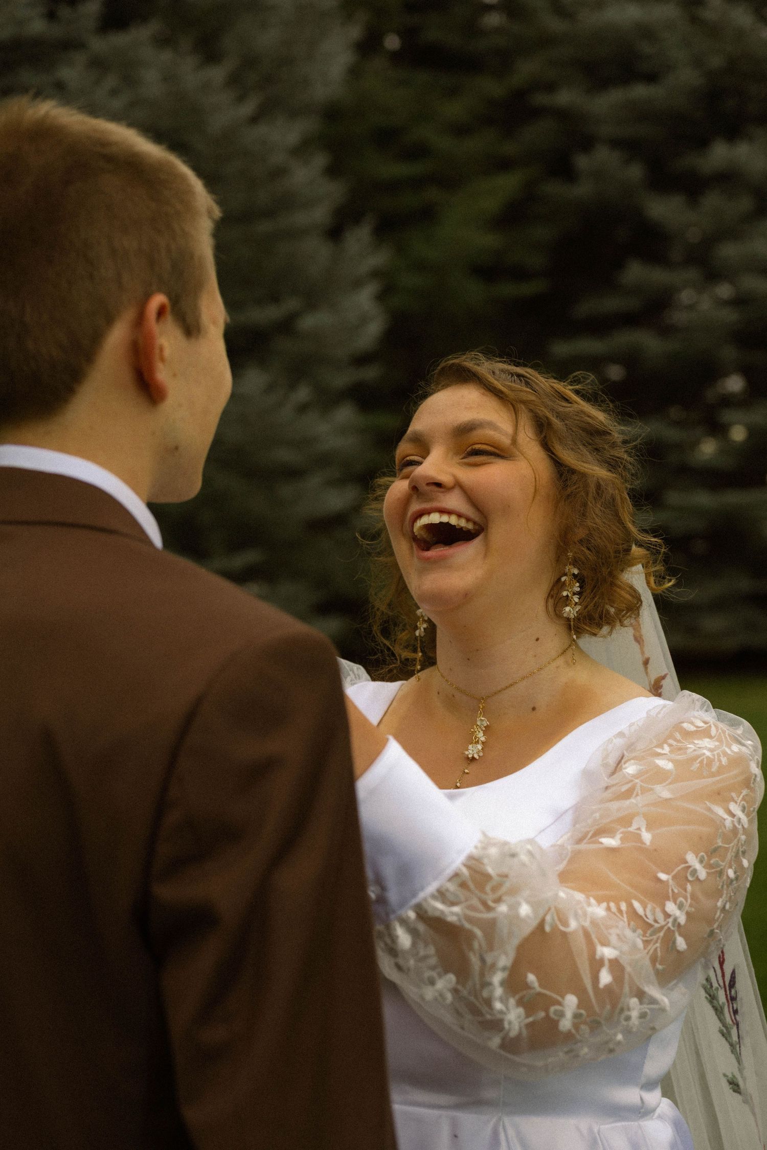Joyful bride in white lace gown laughing with groom in brown suit during outdoor wedding ceremony.