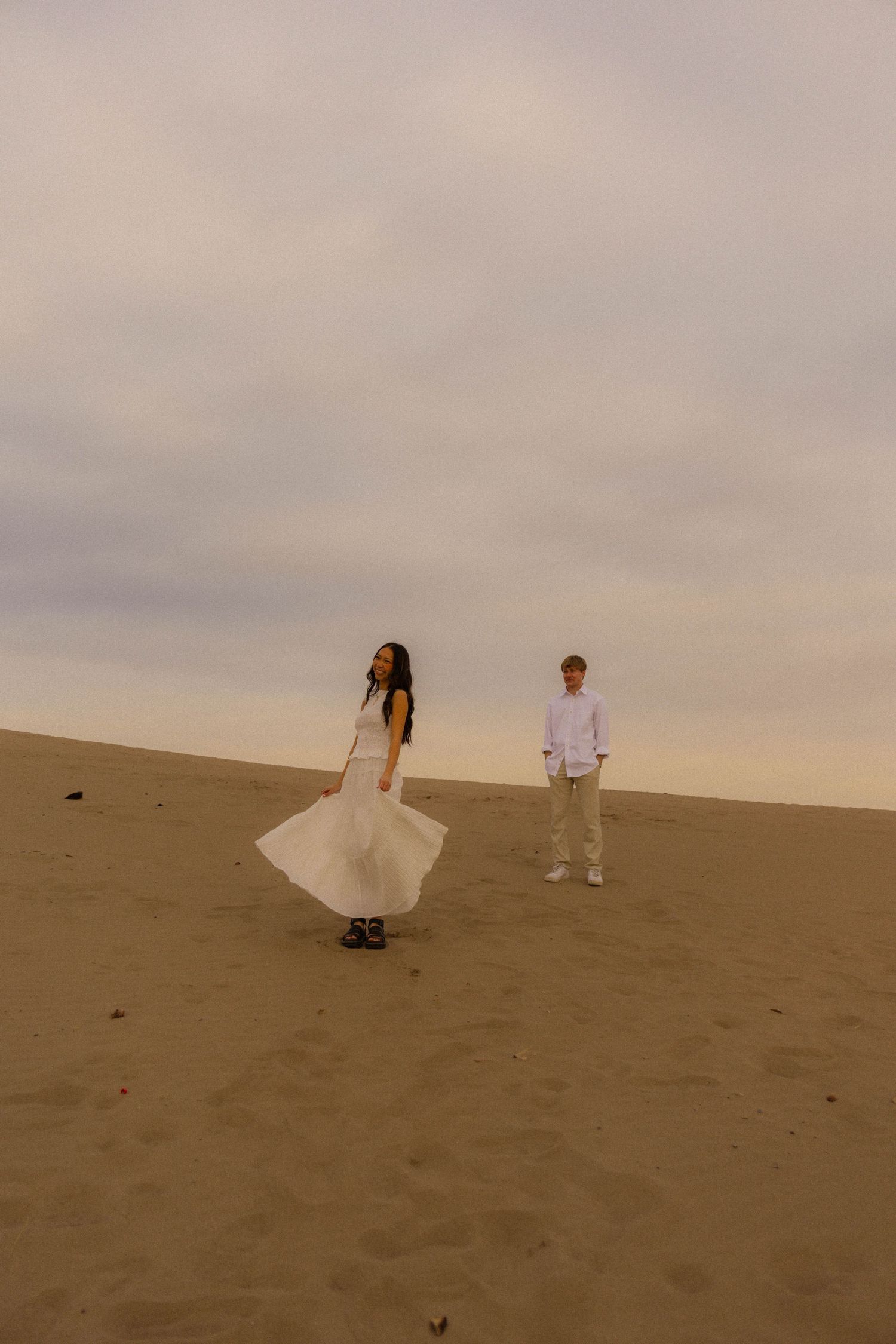 A woman in a white dress and a man in white stand on sandy dunes under a cloudy sky.