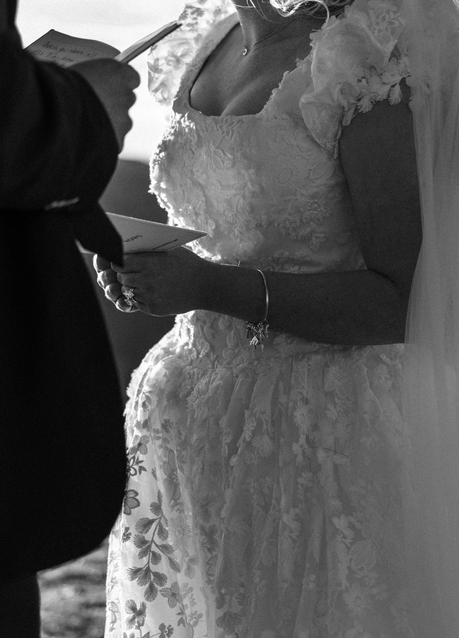 Black and white photo of a bride in lace wedding dress exchanging vows during outdoor ceremony.