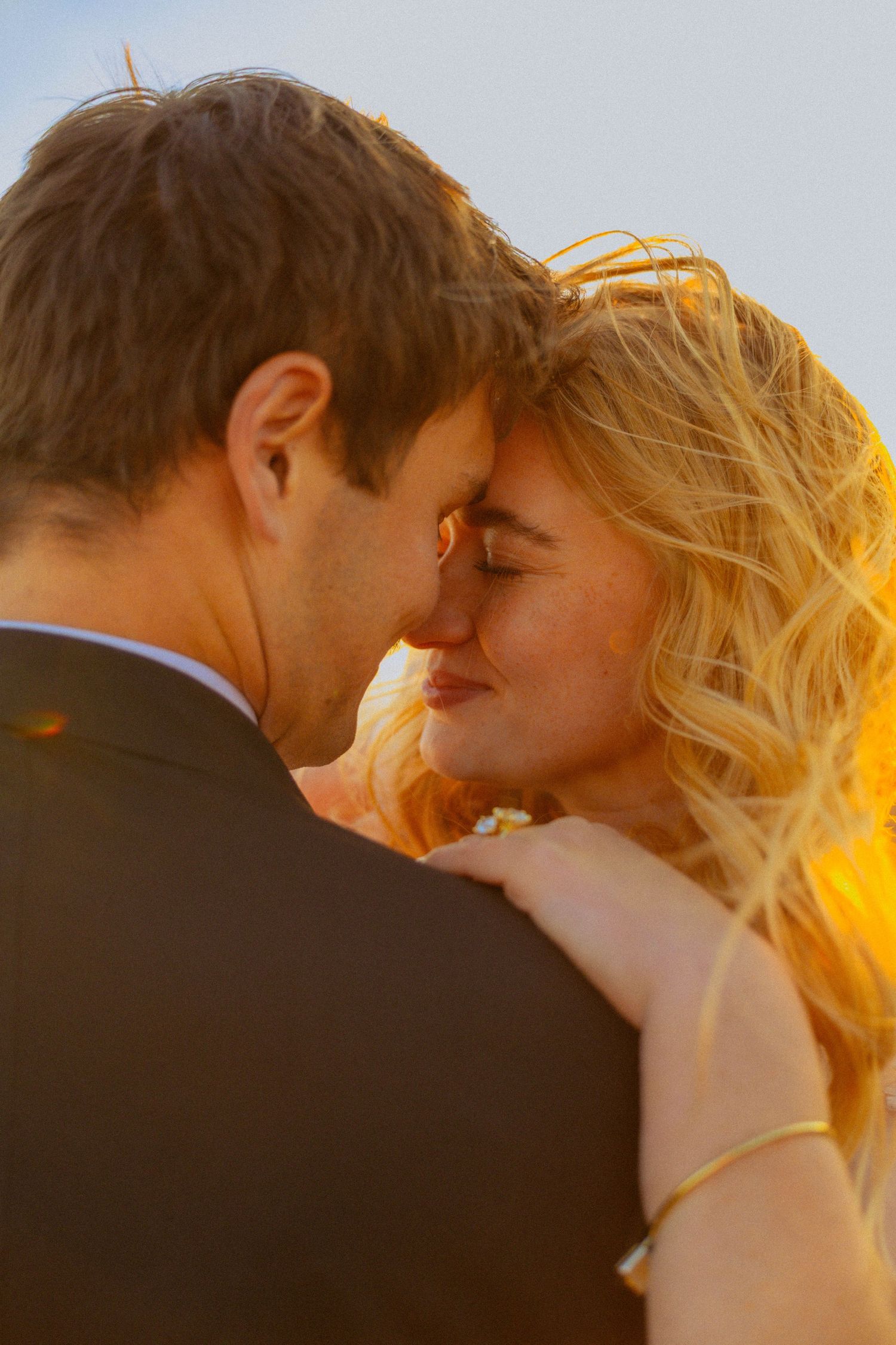 Romantic couple sharing an intimate moment at golden hour sunset, bride with blonde curly hair and groom in suit touching foreheads.