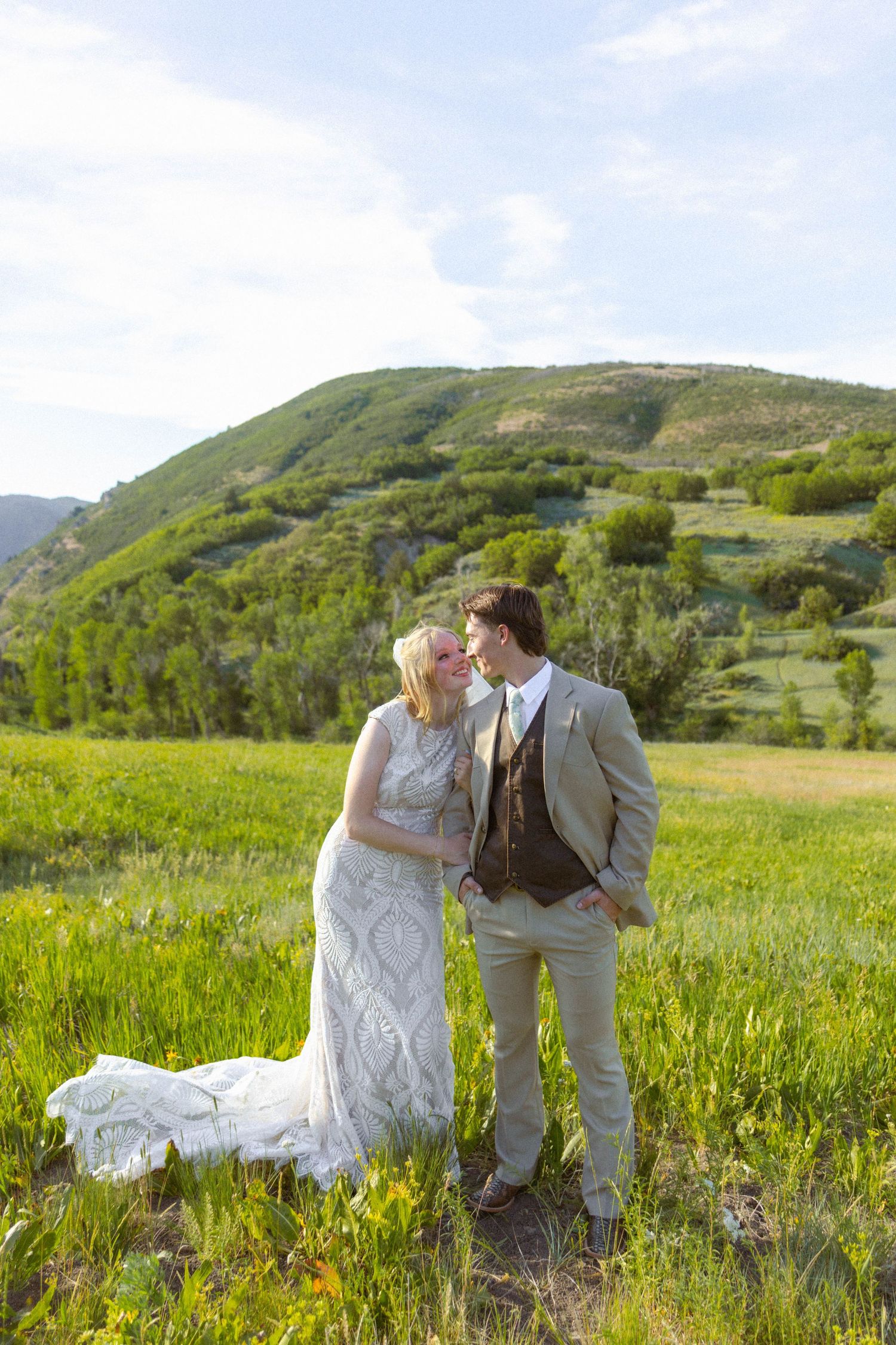 Bride and groom share a romantic moment in a lush green mountain meadow, with the bride in a lace gown.