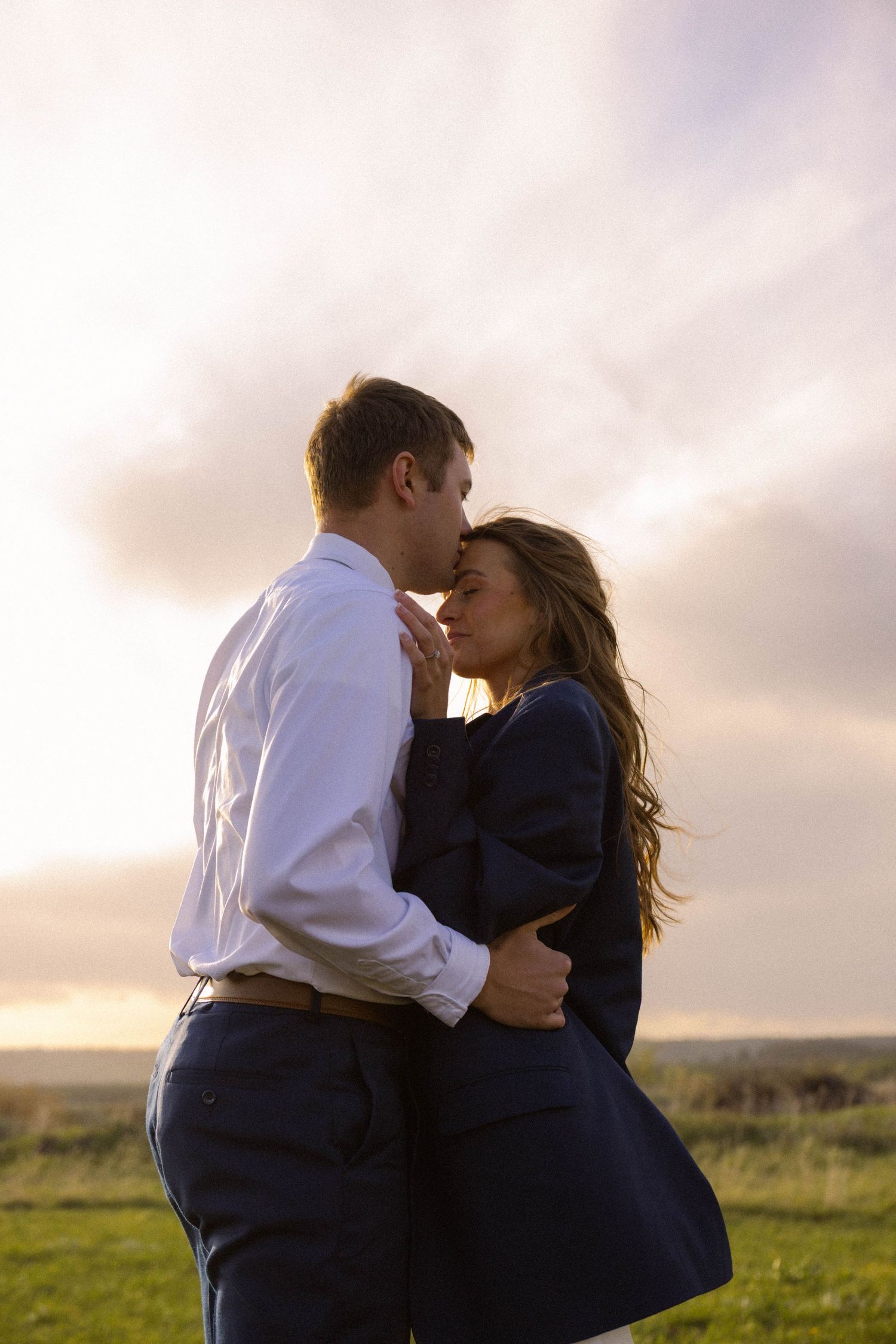 A couple shares a romantic kiss at sunset in a green field, the man in a white shirt and woman in a navy coat.
