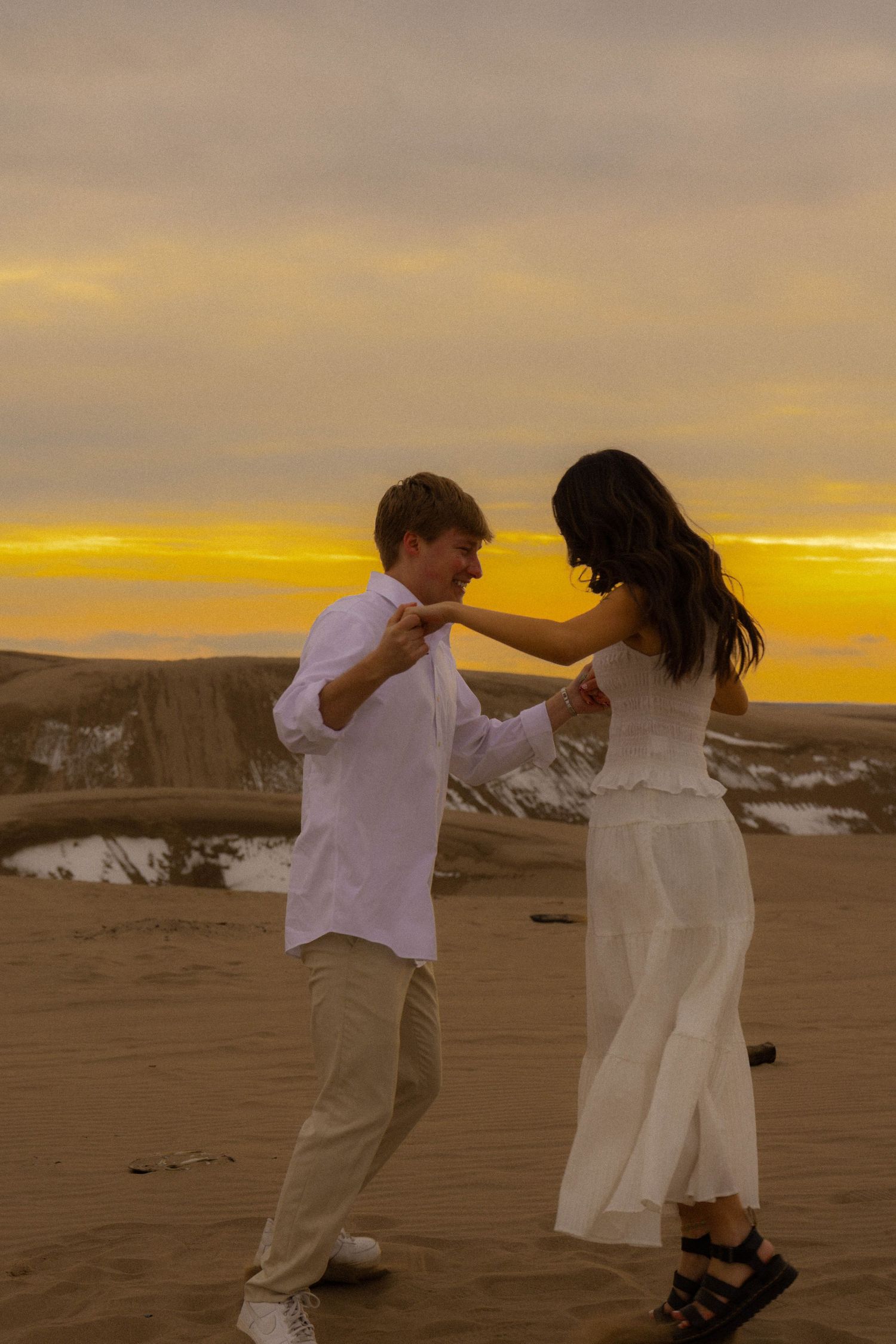 Couple dancing in white outfits on desert sand dunes at golden sunset, romantic outdoor photoshoot.