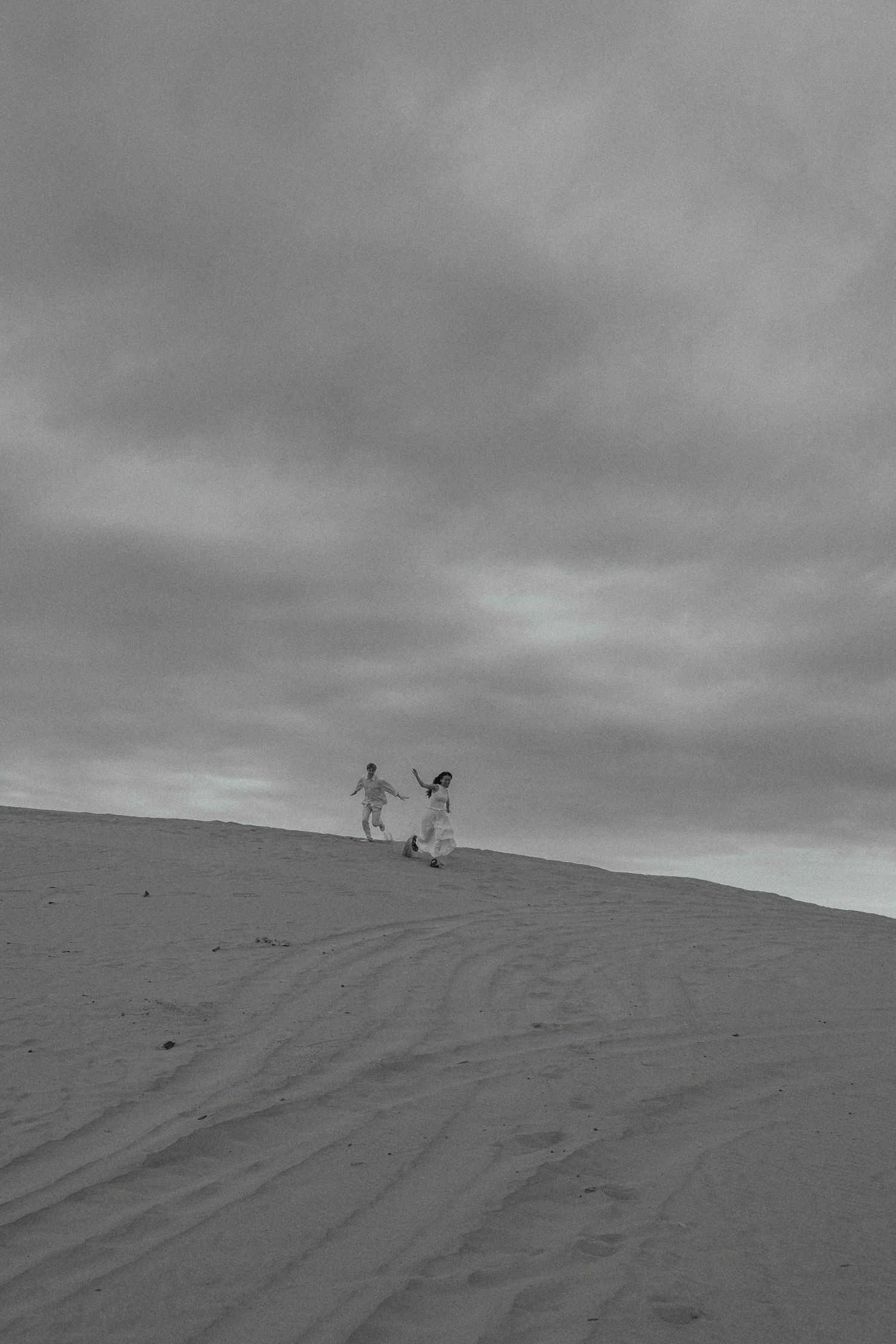 Black and white photo of two people holding hands running on a sand dune under a cloudy sky.