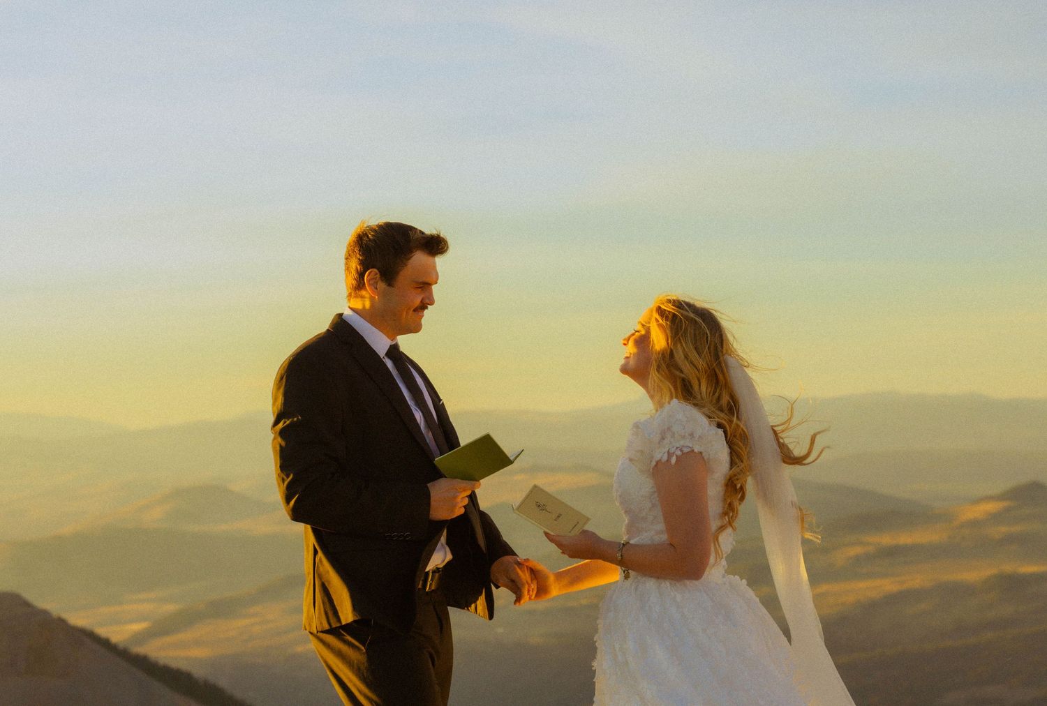 Couple exchanging vows at golden hour on a mountain summit, bride in white lace gown and veil, groom in dark suit.
