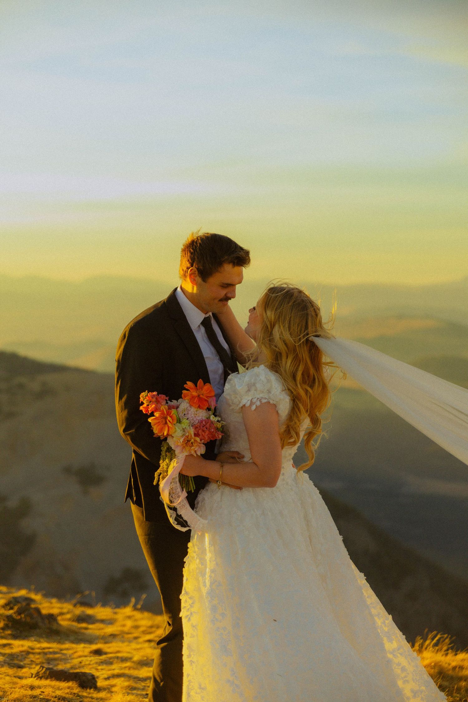 A bride and groom share a romantic moment on a mountain summit at golden hour, her veil flowing in the wind.