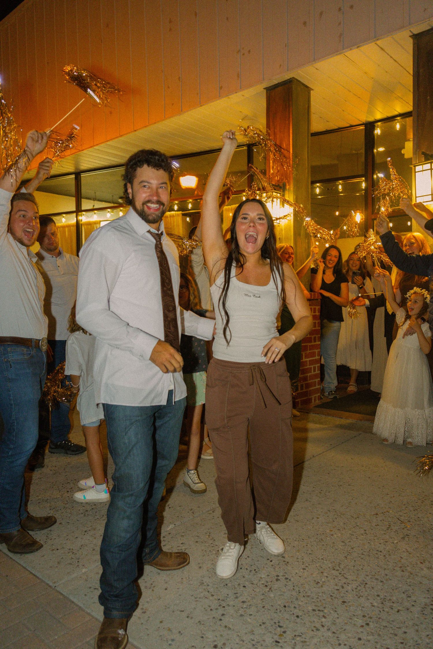 Joyful couple leads celebratory send-off with cheering guests holding sparklers at a festive wedding reception.