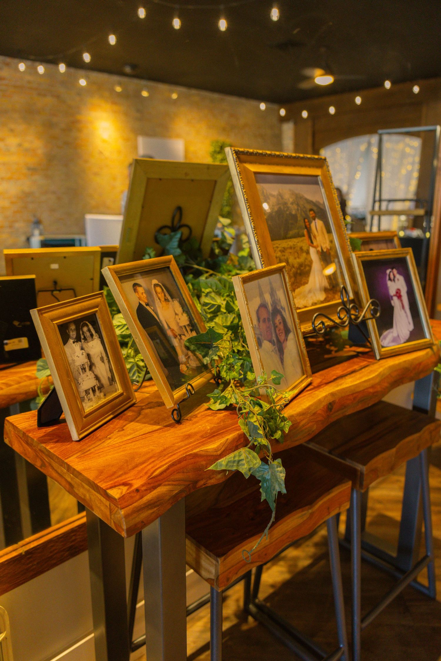 Framed family photos displayed on a rustic wooden table with ivy greenery, set in a warmly lit venue with string lights.