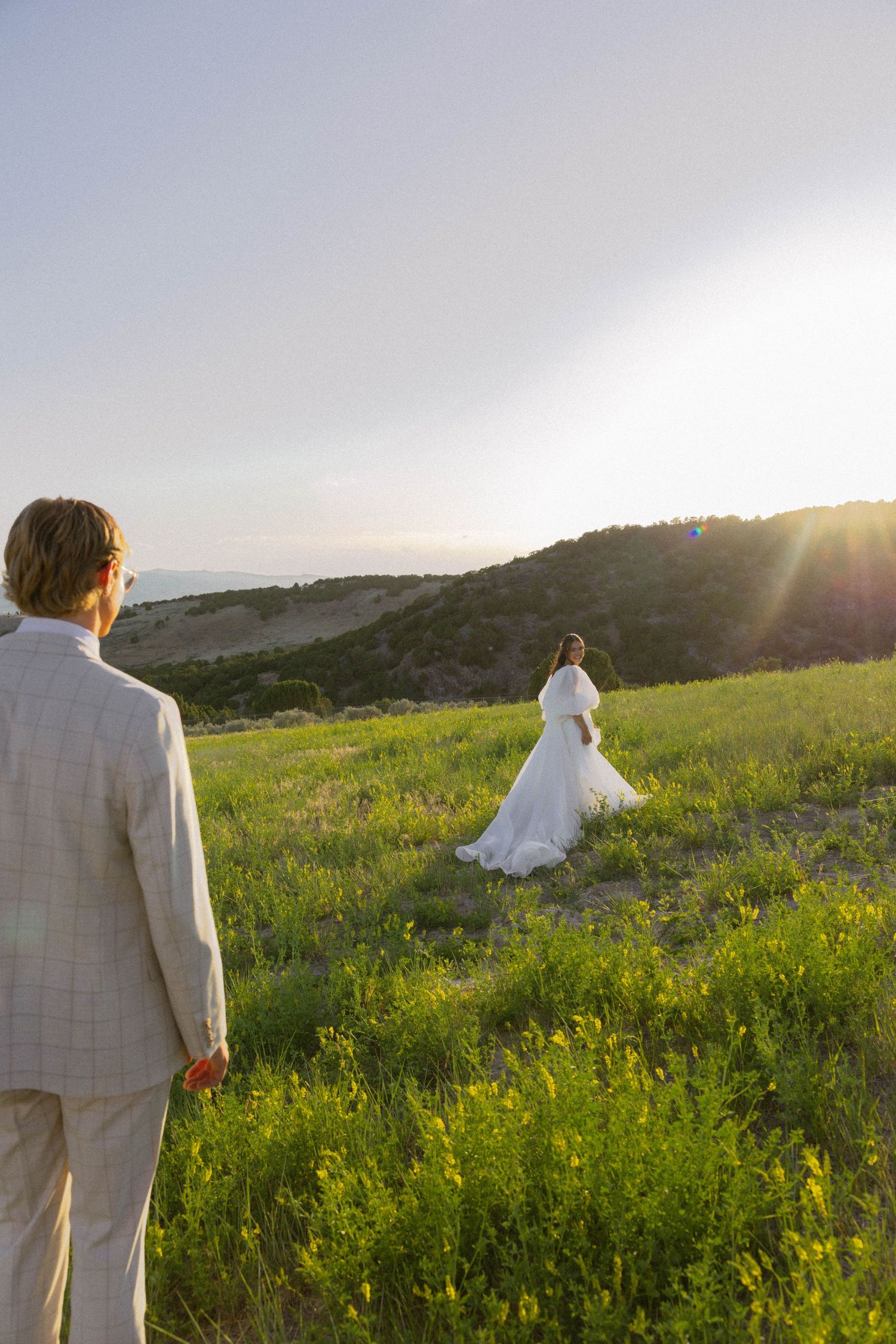 A bride in a white gown walks through a sunlit green meadow as a groom in a tan suit watches during golden hour.