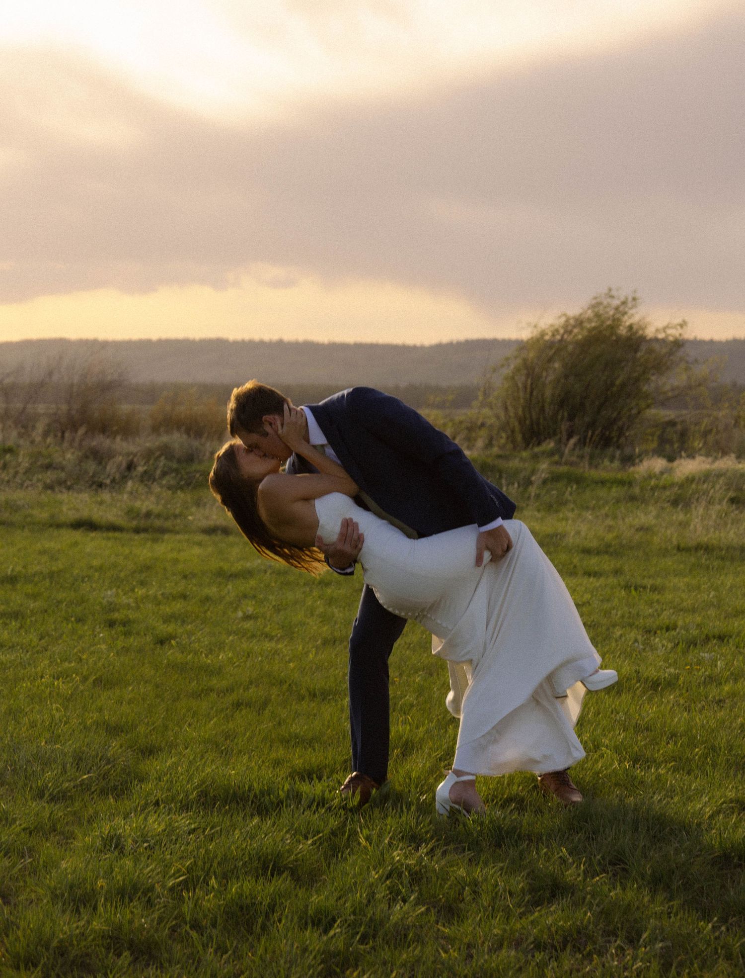 A groom dips his bride for a romantic kiss in a lush green field at golden hour during their wedding.