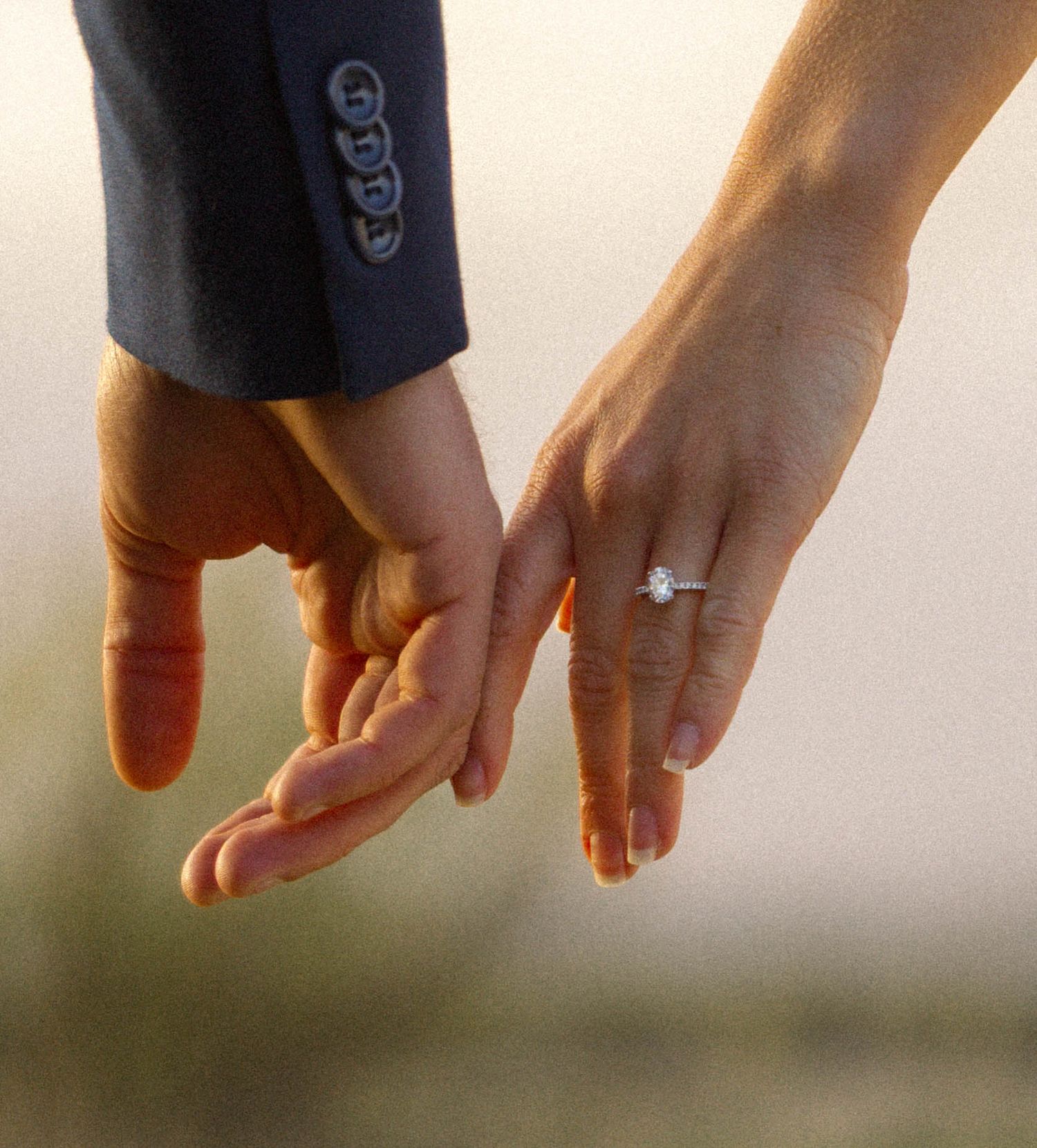 Close-up of two hands gently touching, woman wearing a diamond engagement ring, warm golden sunset light.