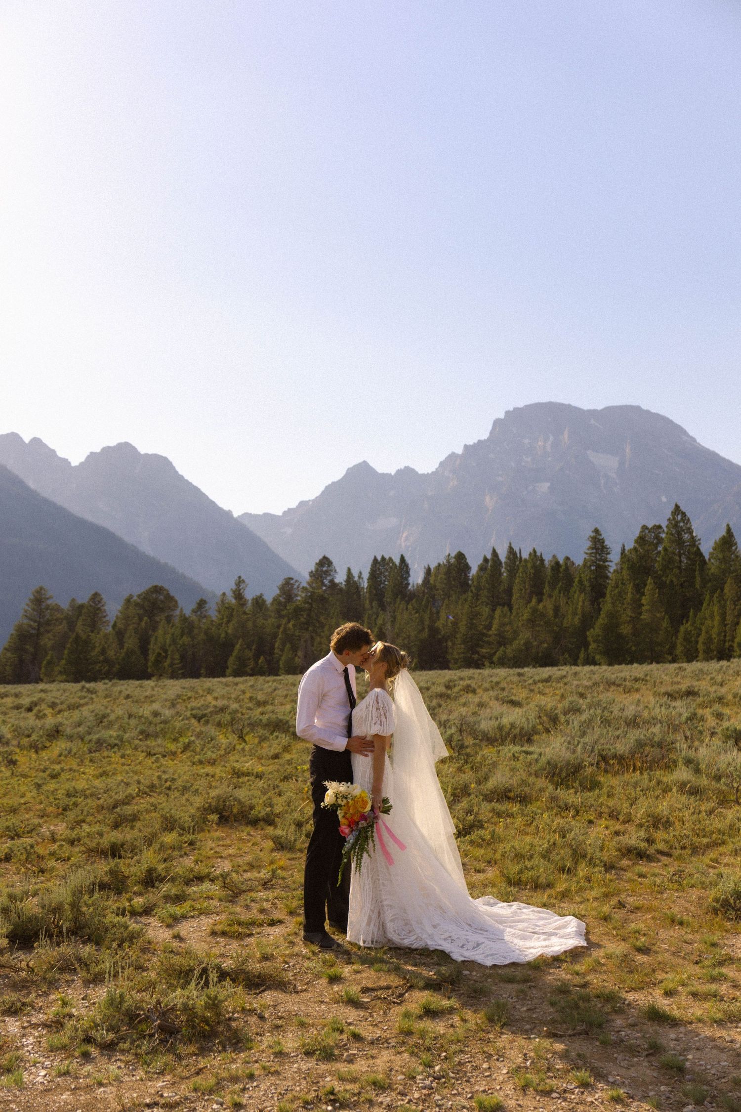 Bride and groom share a romantic kiss in a mountain meadow with Grand Teton peaks and pine trees in the background.