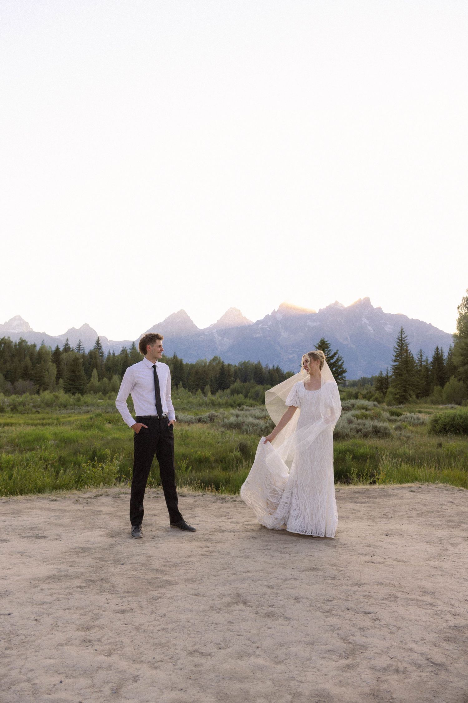 Bride and groom stand apart in mountain meadow, her white gown and veil flowing in breeze, Grand Tetons backdrop.