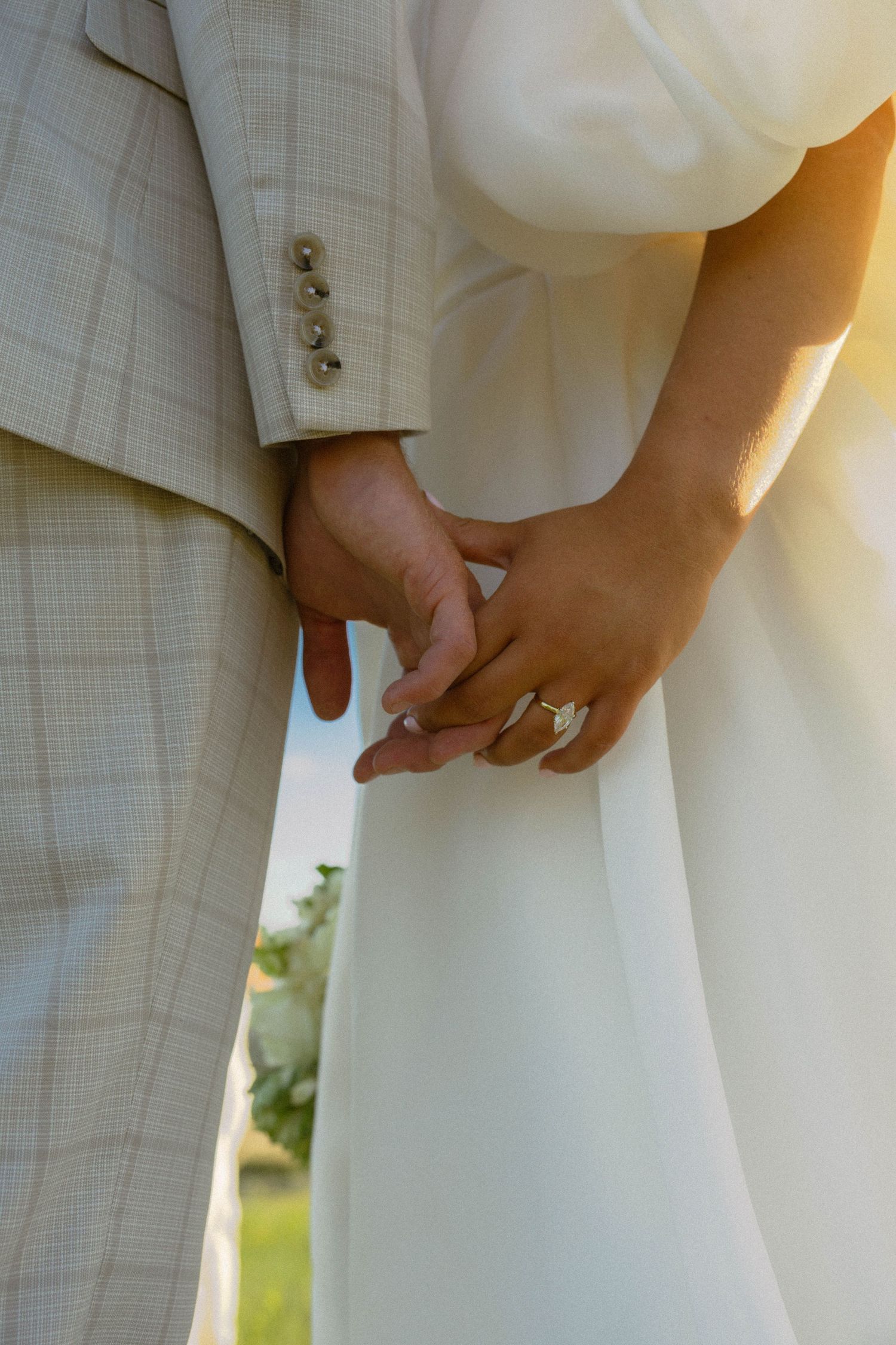 Bride and groom holding hands at golden hour, showing wedding ring on bride's finger, wearing white gown and beige suit.