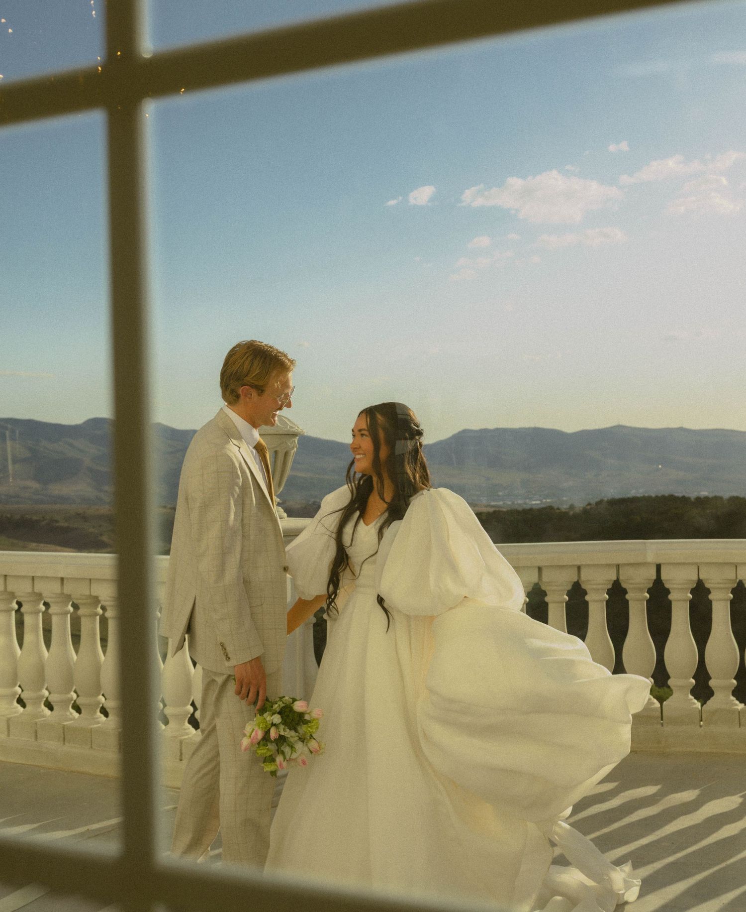 Bride and groom share a romantic moment on a balcony with mountain views, framed by a window.